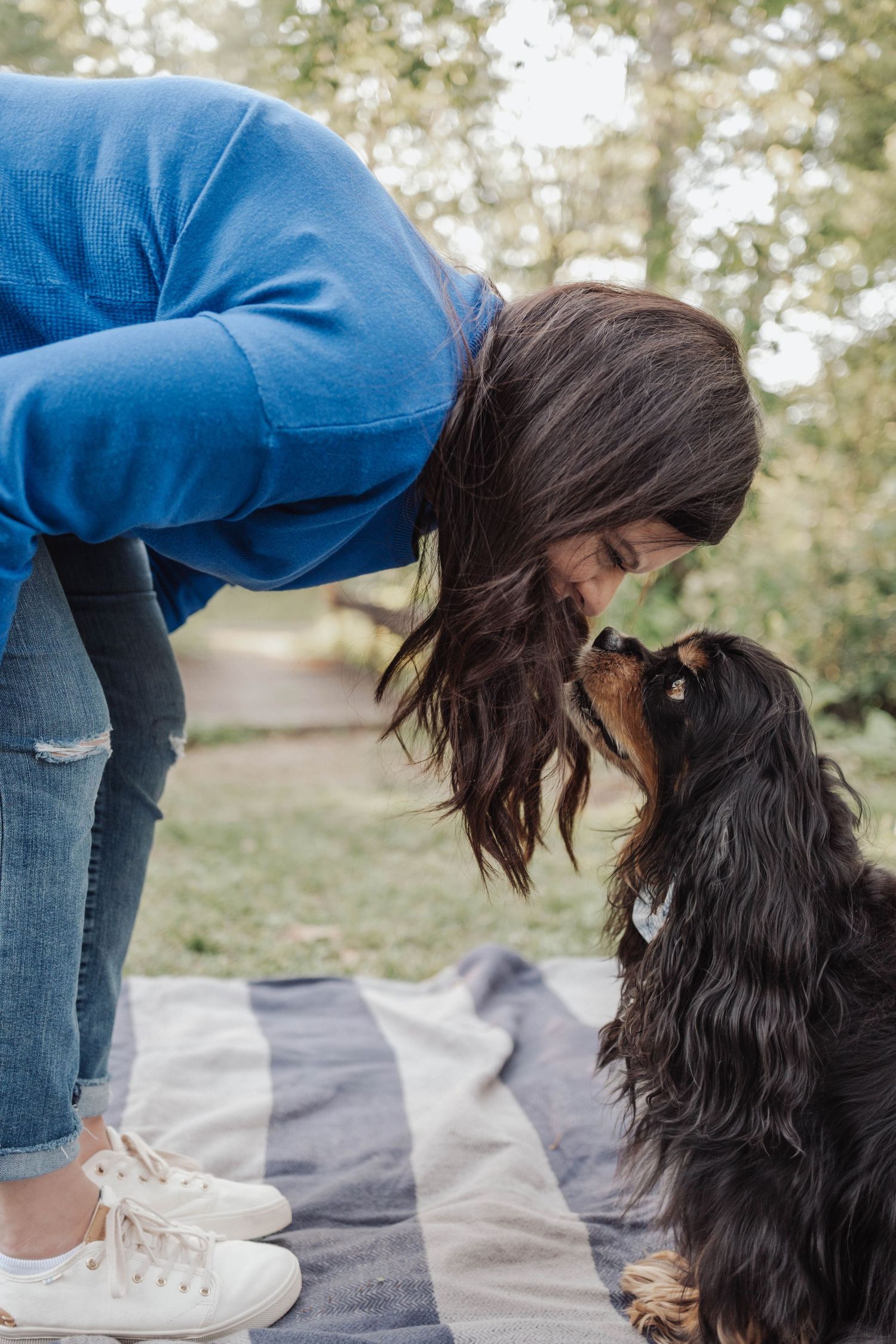 Woman in blue sweater leaning down to kiss her Cavalier King Charles Spaniel on a striped blanket outdoors.