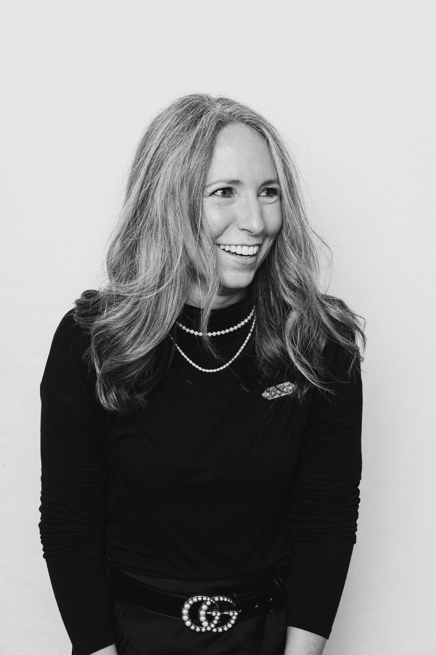 Smiling woman with long hair wearing black turtleneck, pearl necklace, and Gucci belt in black and white photo.