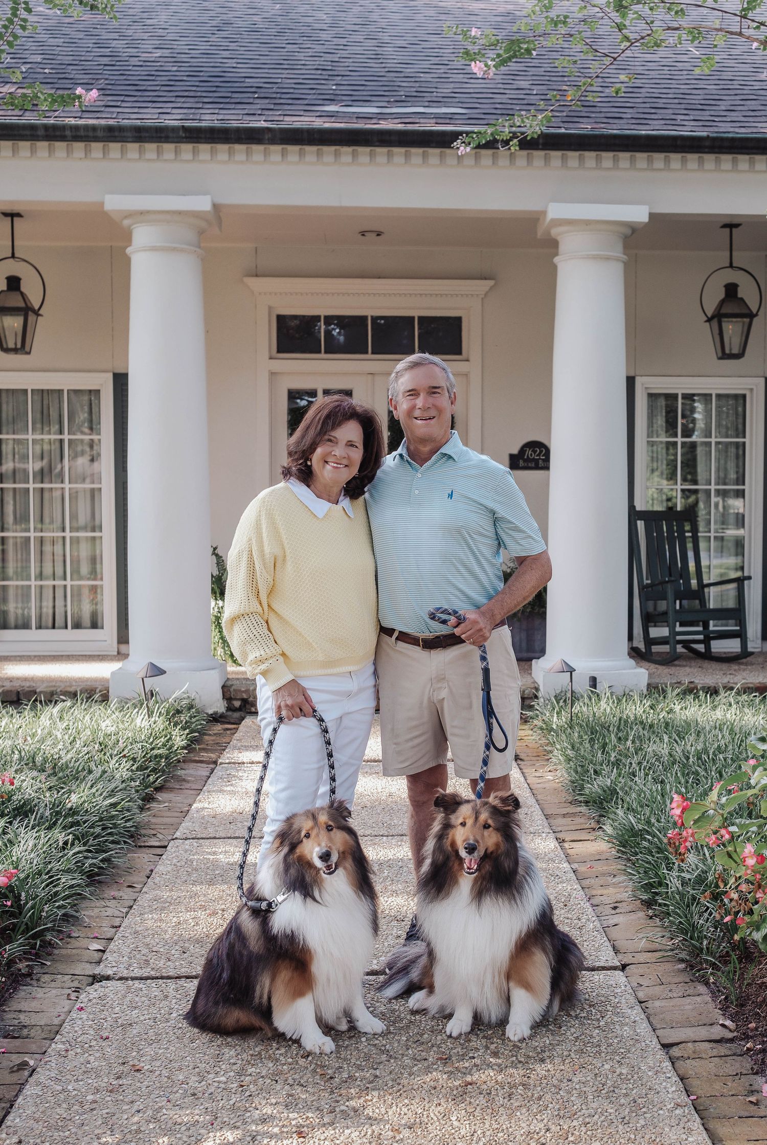 Smiling couple posing with two Shetland Sheepdogs on a brick walkway in front of a white columned home.