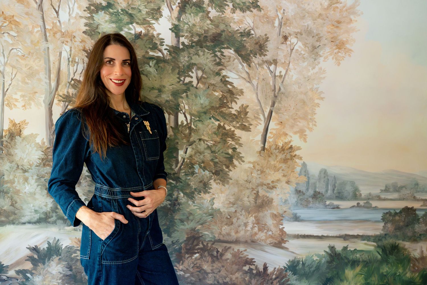 Smiling woman with long brown hair in a denim jumpsuit stands with a hand in her pocket before a hand-painted scenic landscape mural.