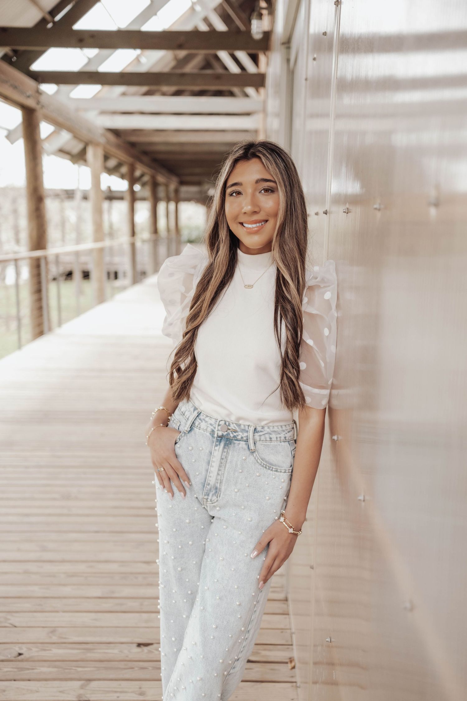 Young woman smiling in white sheer-sleeve top and embellished jeans, leaning against metal wall on wooden walkway.