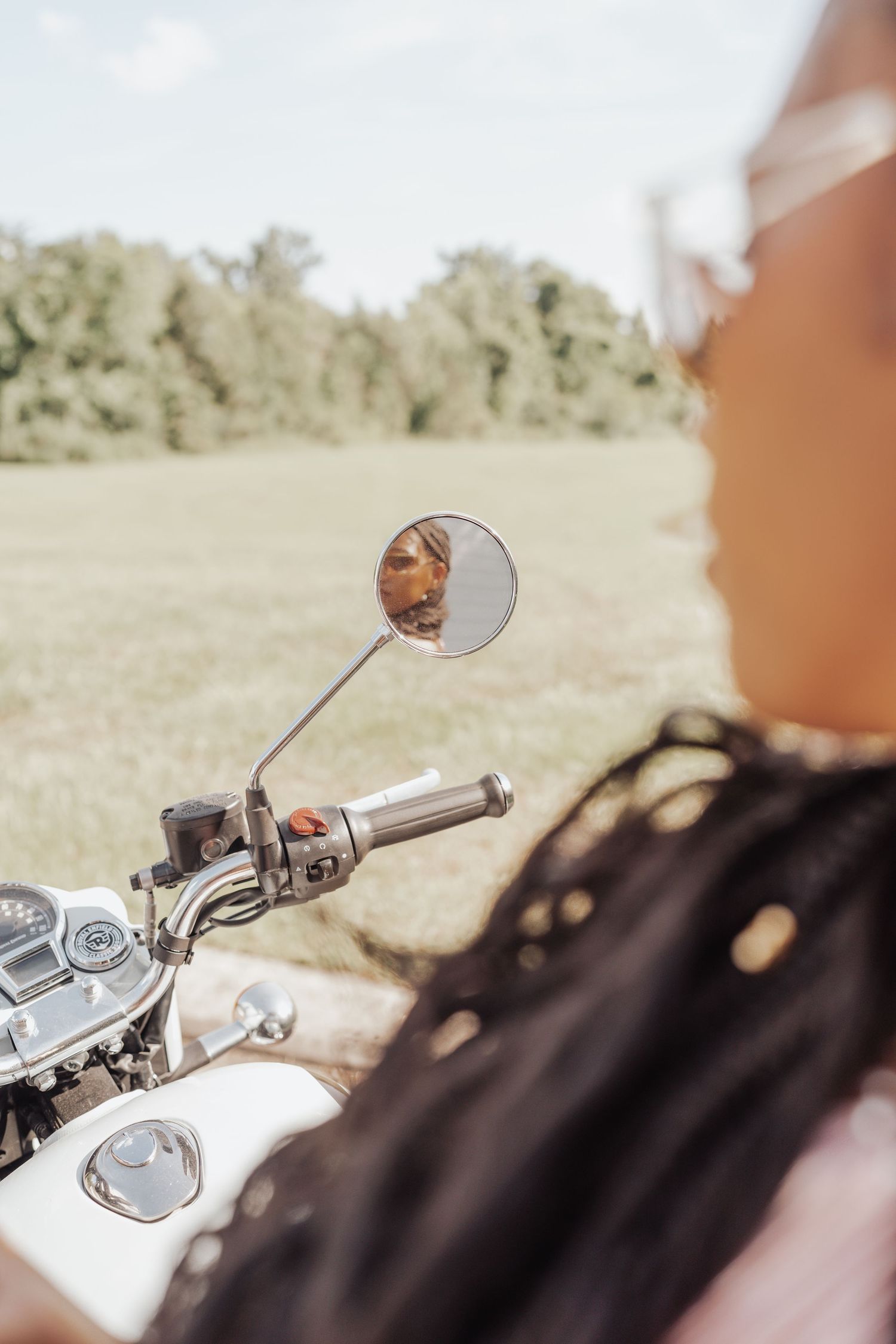 Woman with long braids on motorcycle viewed through round side mirror reflecting her face outdoors.