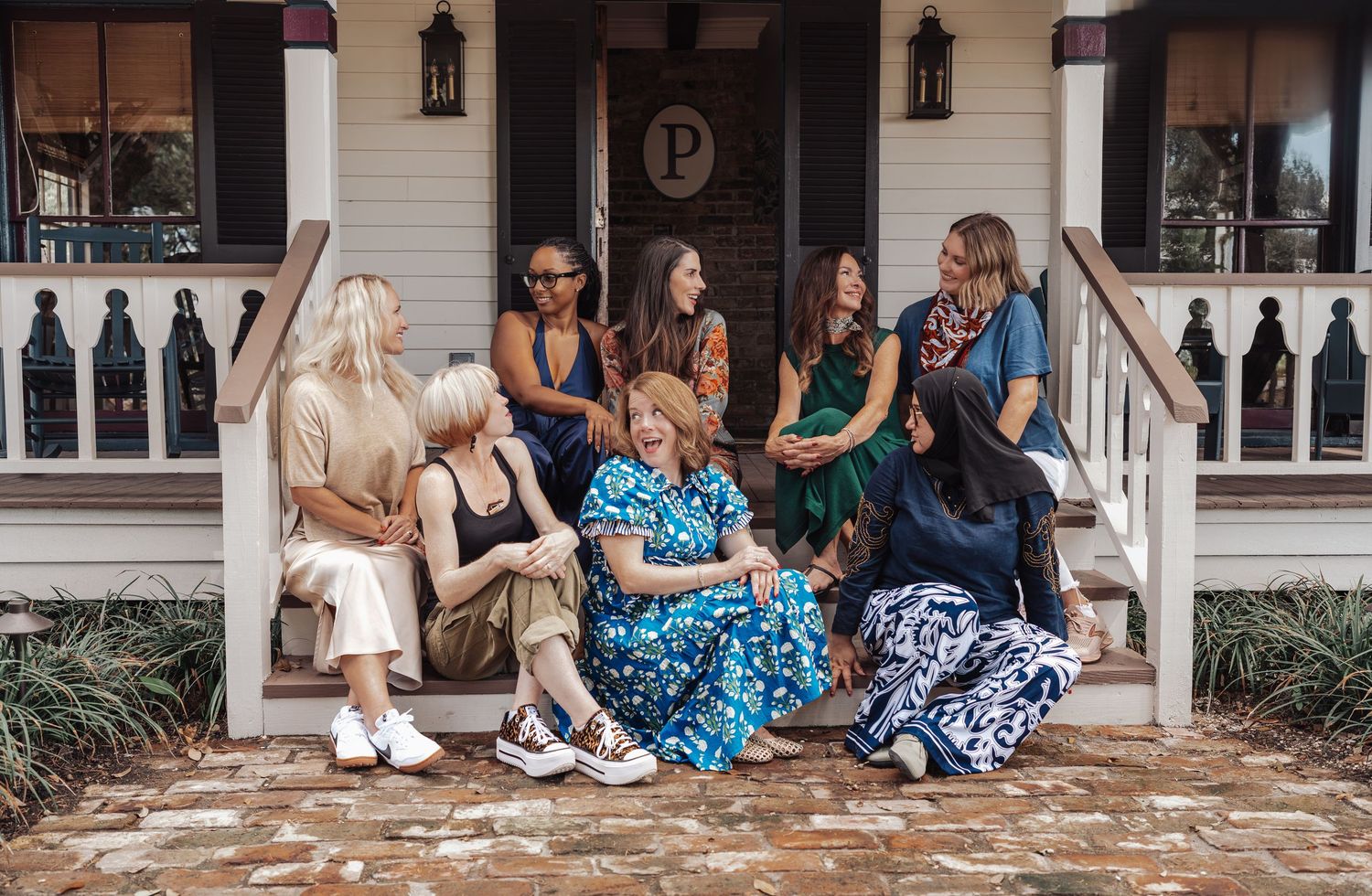 Group of smiling women in colorful patterned outfits sit together on the front porch steps of a white house with a dark wooden door.