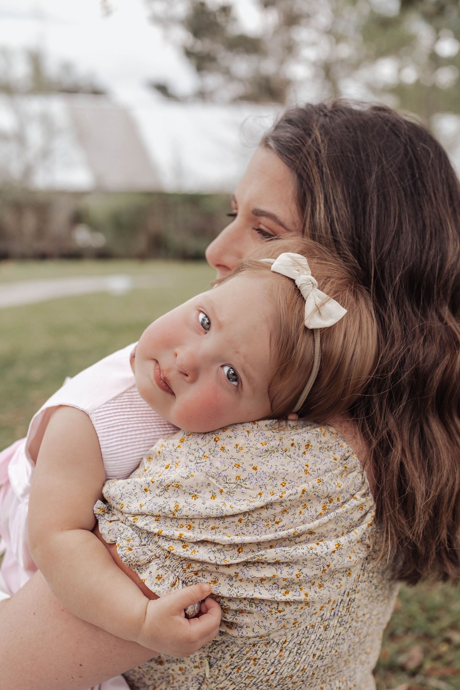 Mother holding baby girl with bow headband, sharing a tender embrace outdoors in a garden setting.