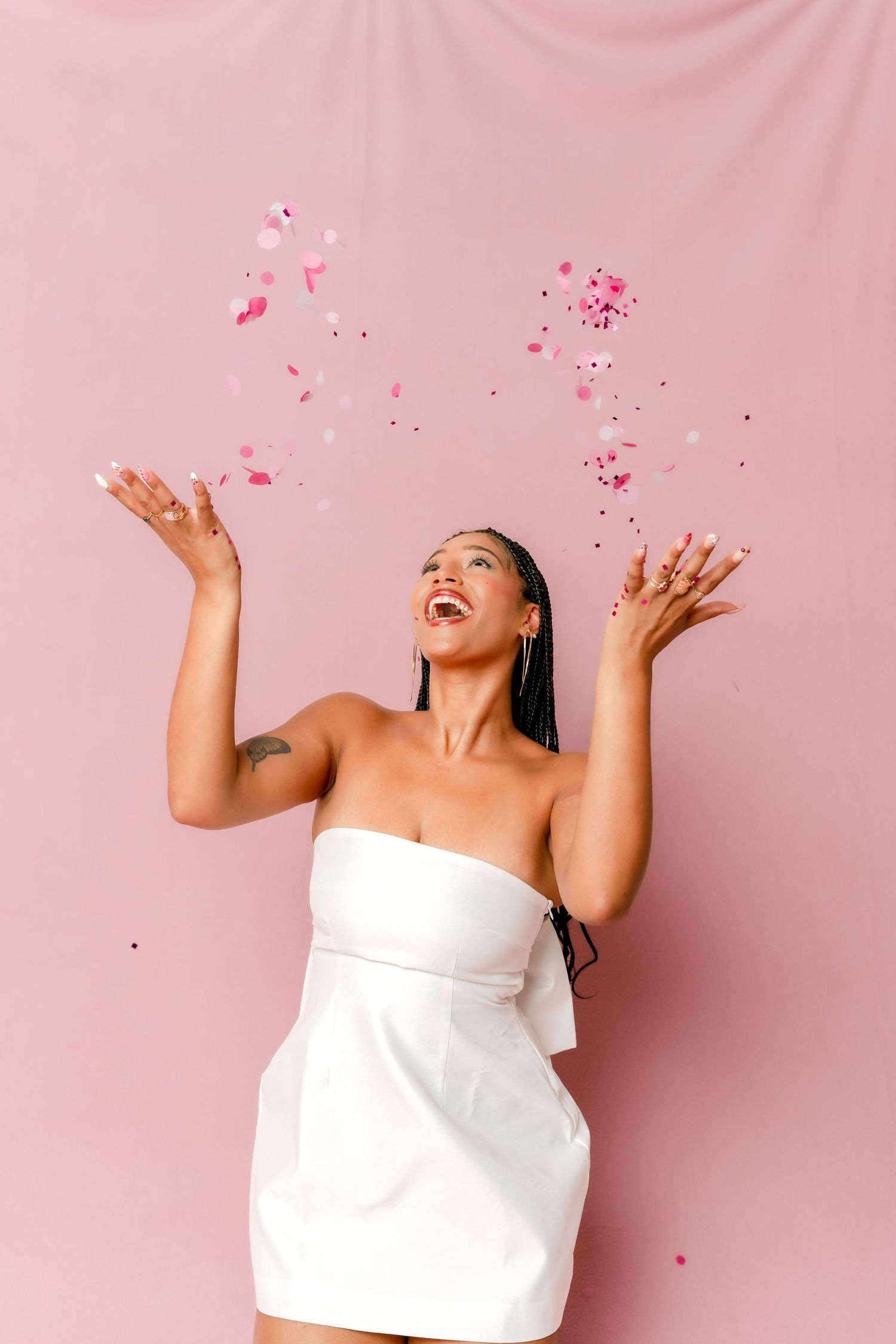 Joyful woman in white strapless dress tossing pink confetti in the air against pink background.