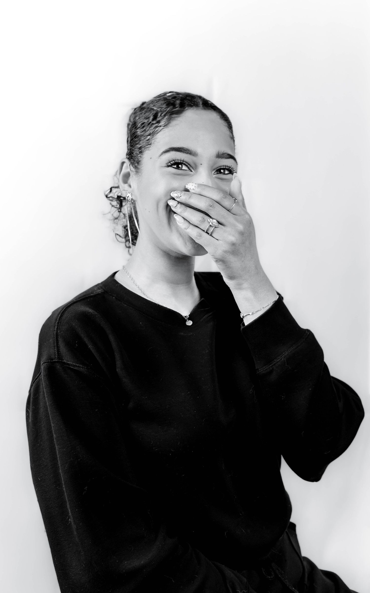 Black and white portrait of a laughing young woman in black sweater covering her mouth with her hand, wearing hoop earrings.