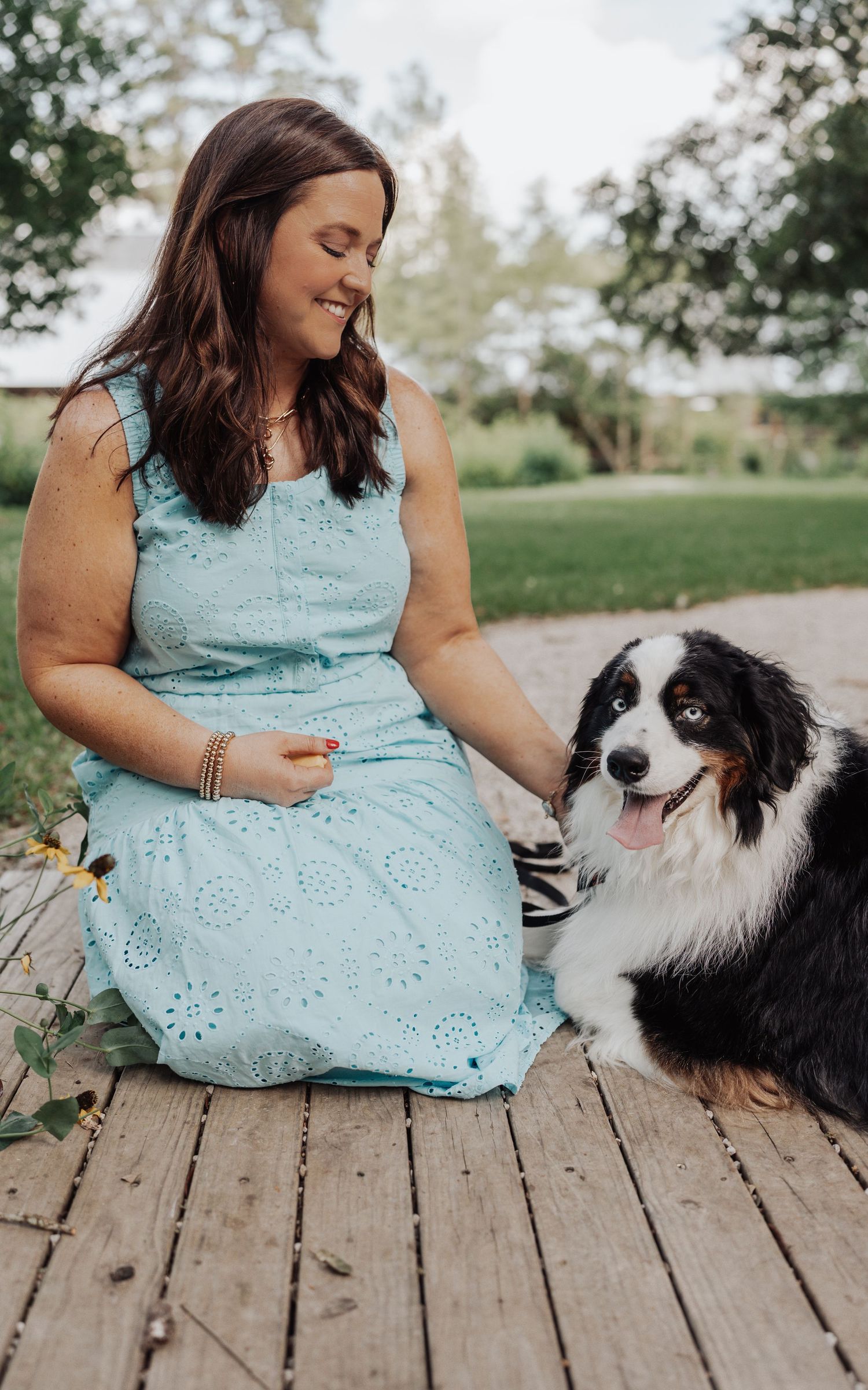 Smiling woman in light blue eyelet dress sitting on wooden deck with black and white Australian Shepherd dog outdoors.