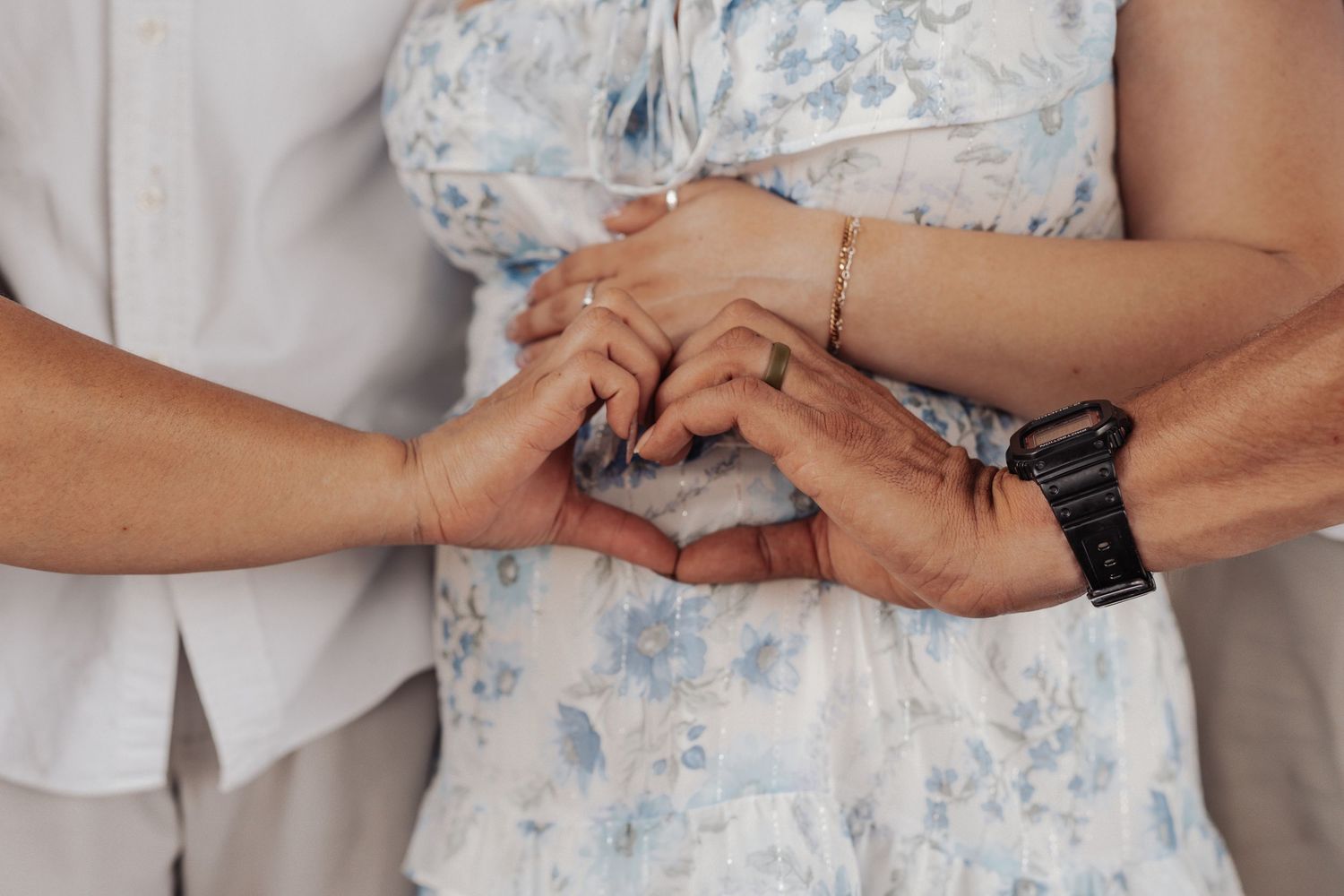 Three people forming a heart shape with their hands, wearing casual and floral outfits, symbolizing love and togetherness.
