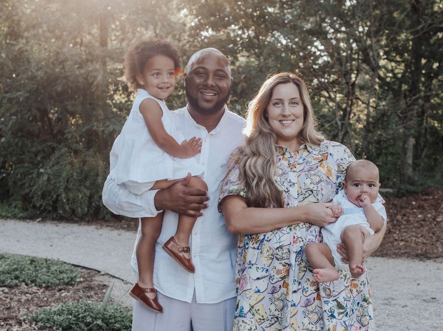 A family wearing coordinated white and floral outfits poses together outdoors in a natural setting during golden hour.