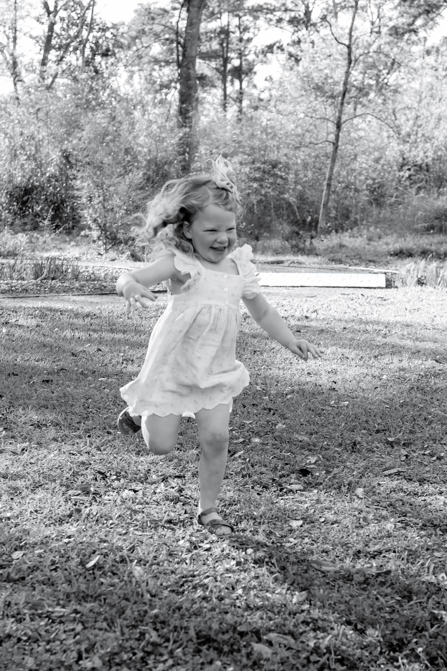 A happy toddler girl in a white dress runs barefoot through a grassy yard in this black and white photo.