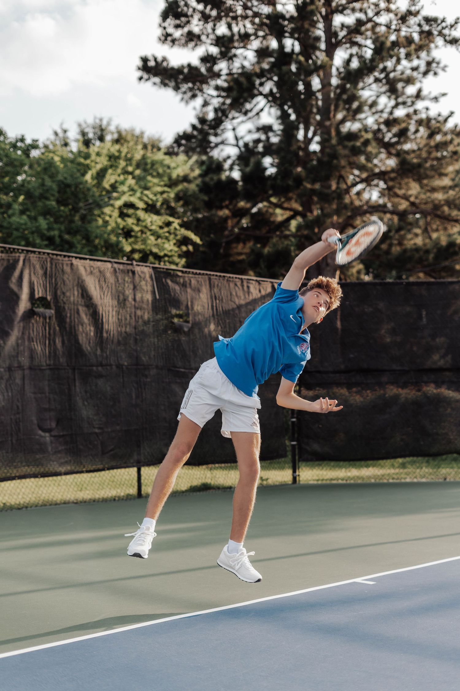 Young male tennis player in blue shirt executing a powerful serve on an outdoor hard court.