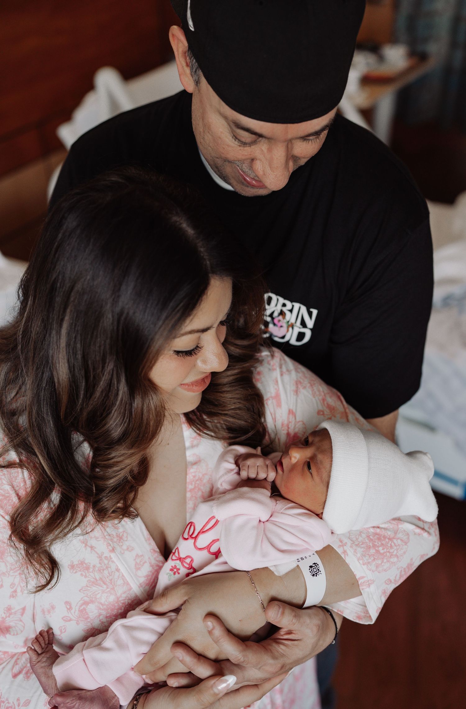 Smiling new parents in a hospital room gently cradle their sleeping newborn baby dressed in a pink outfit and white knit hat.