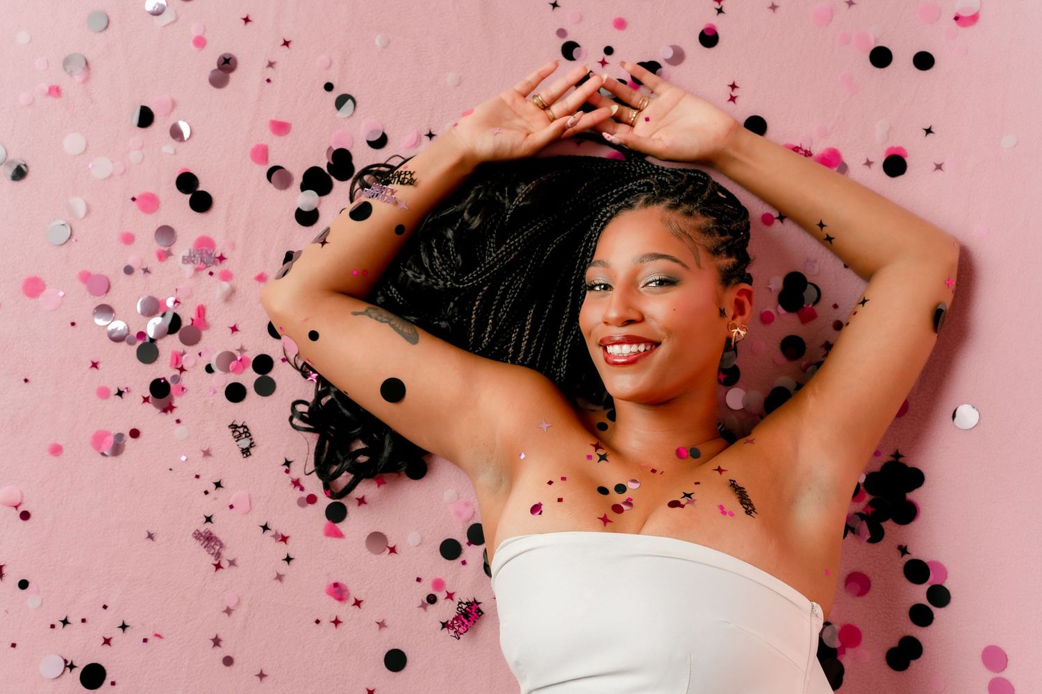 Young Black woman smiling, lying on pink background with colorful confetti, wearing white strapless top.