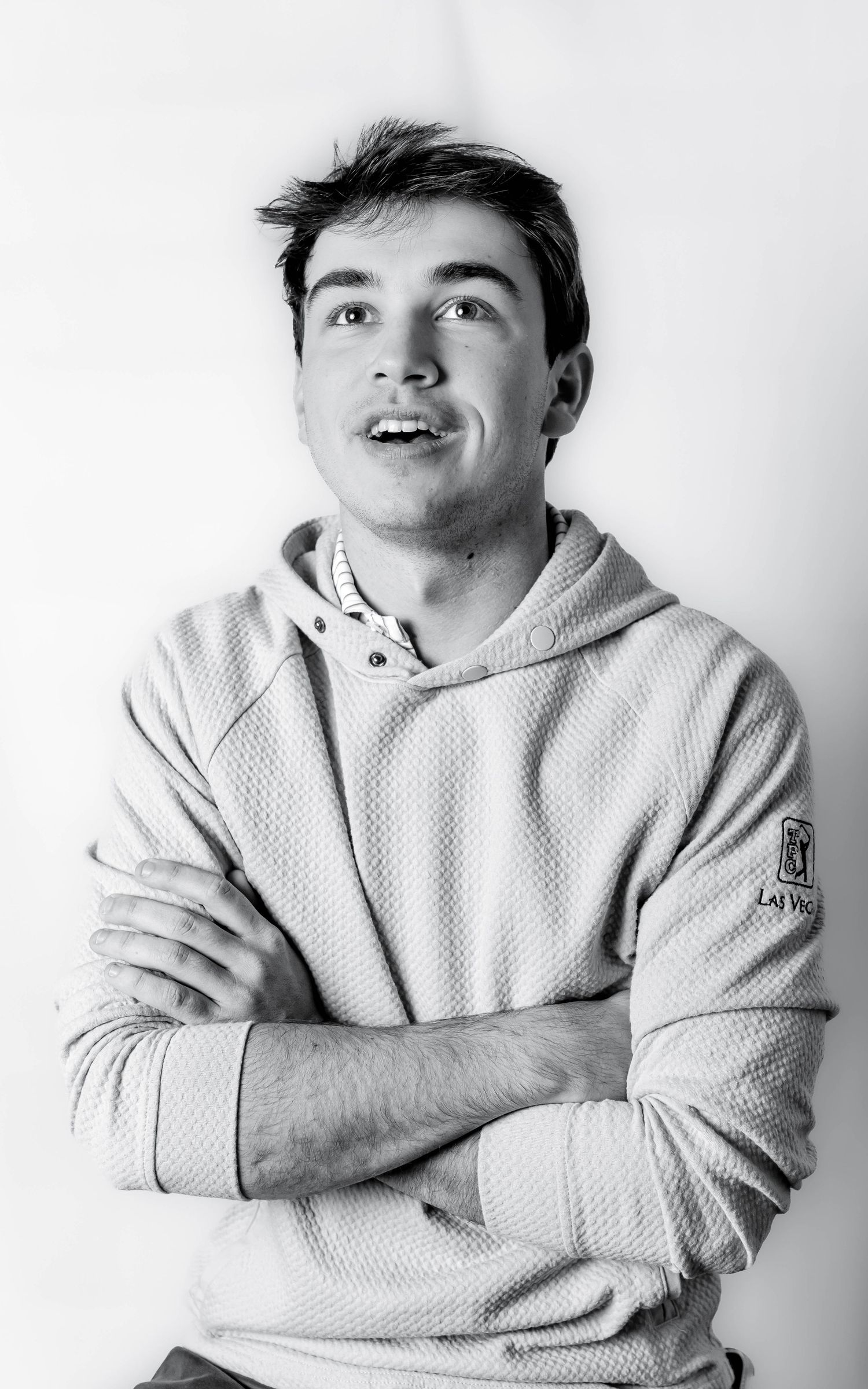 Black and white portrait of a young man in a hoodie, arms crossed, looking up with a confident smile.