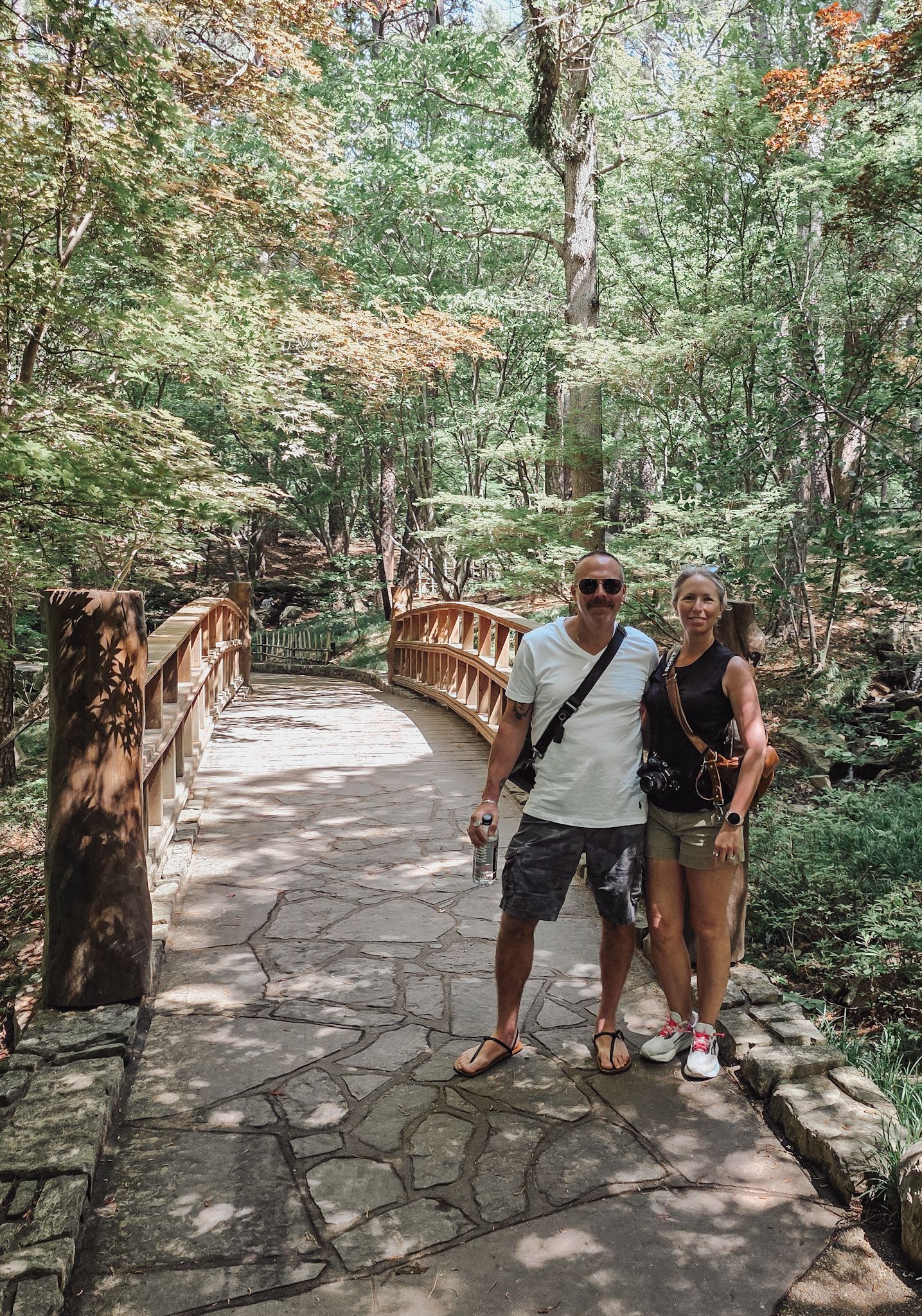 Couple posing on a stone path wooden bridge surrounded by lush green trees in a forest park.