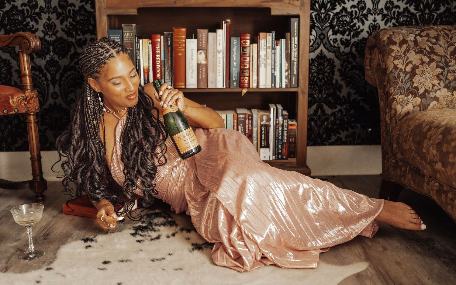 Woman in pink satin dress lounging on floor holding champagne bottle near bookshelf in elegant room.
