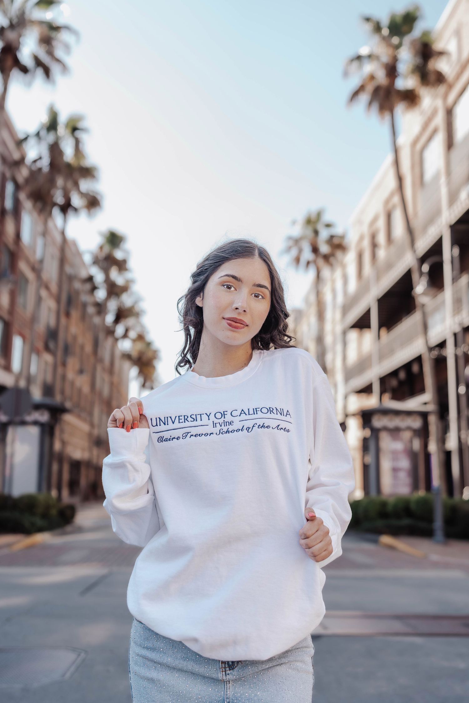 Young woman in University of California white sweatshirt poses on palm tree-lined street.