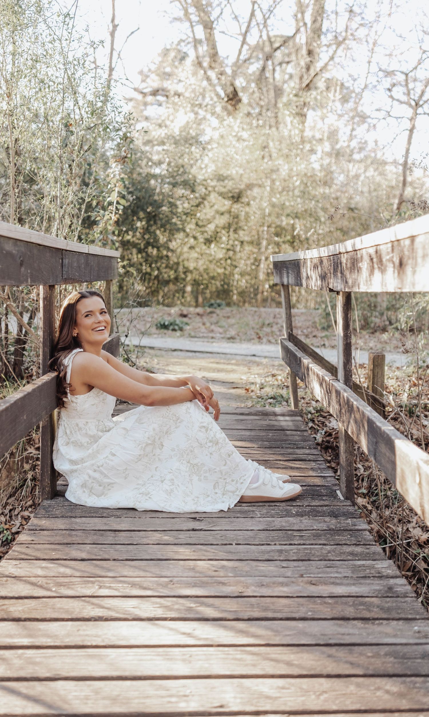 A person in a white wedding dress poses playfully on a rustic wooden bridge in a natural outdoor setting.