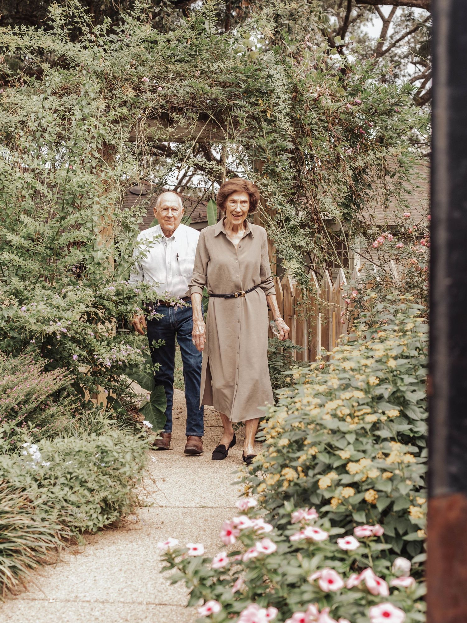 People walking along a gravel garden path bordered by lush greenery and blooming pink flowers.