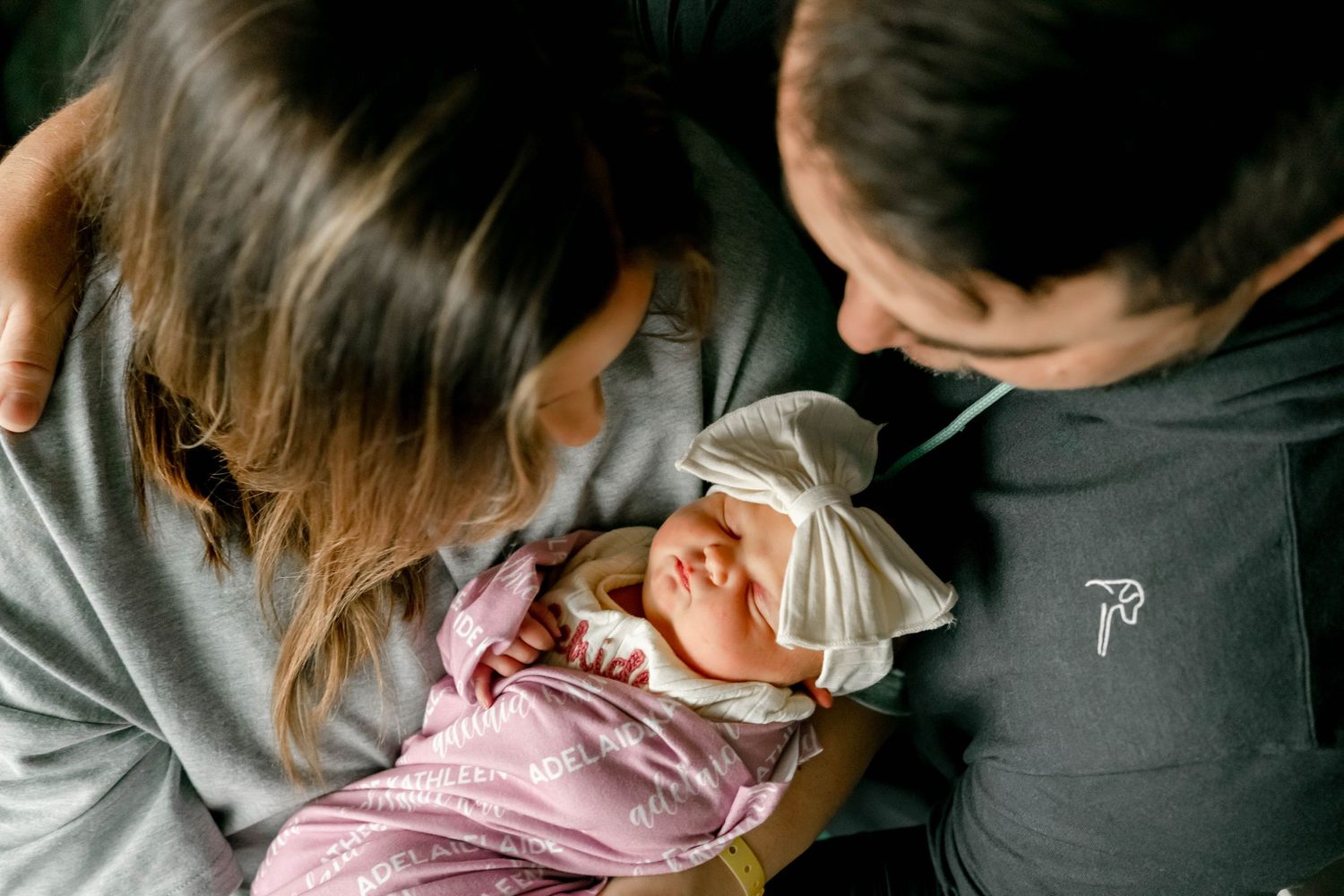 Two parents lovingly gazing at their newborn baby wrapped in a pink swaddle with a white bow headband.