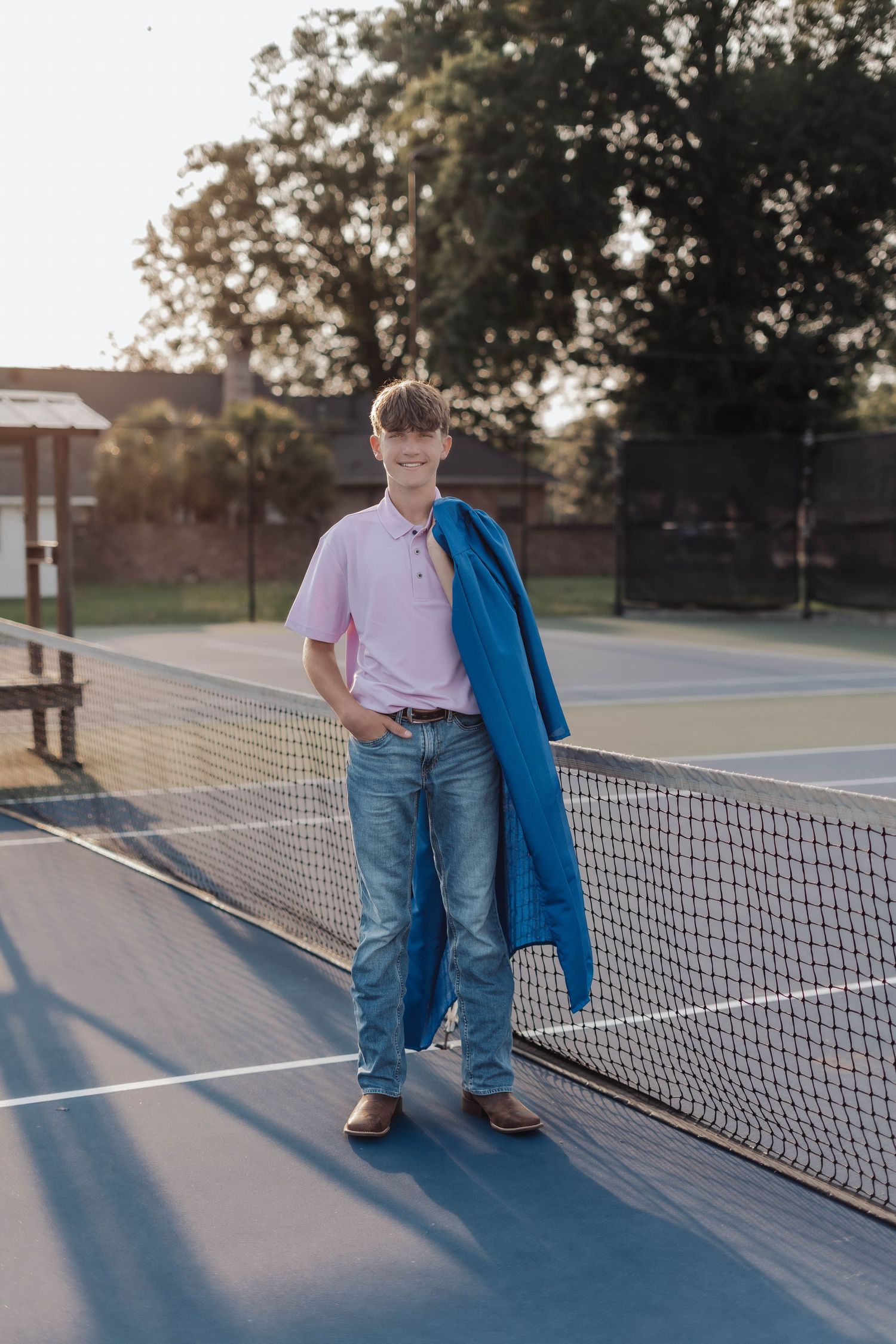 Teen boy in pink polo and jeans poses by tennis net holding blue jacket on sunny outdoor court.