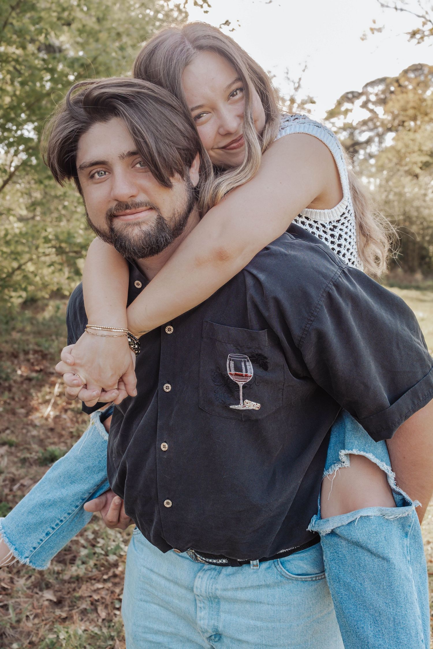 A couple shares a joyful piggyback ride outdoors, the woman hugging the bearded man wearing a black shirt.