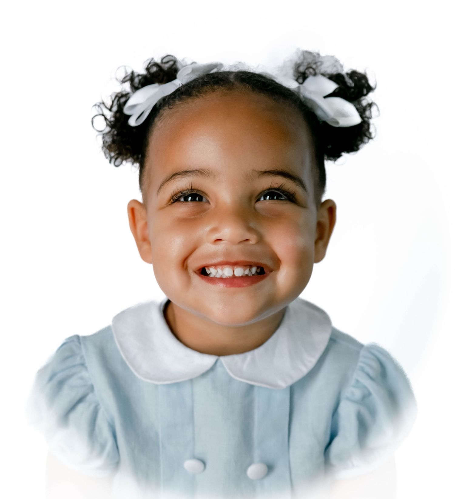 Adorable young child with curly hair and a bright smile wearing a white dress against a light background.