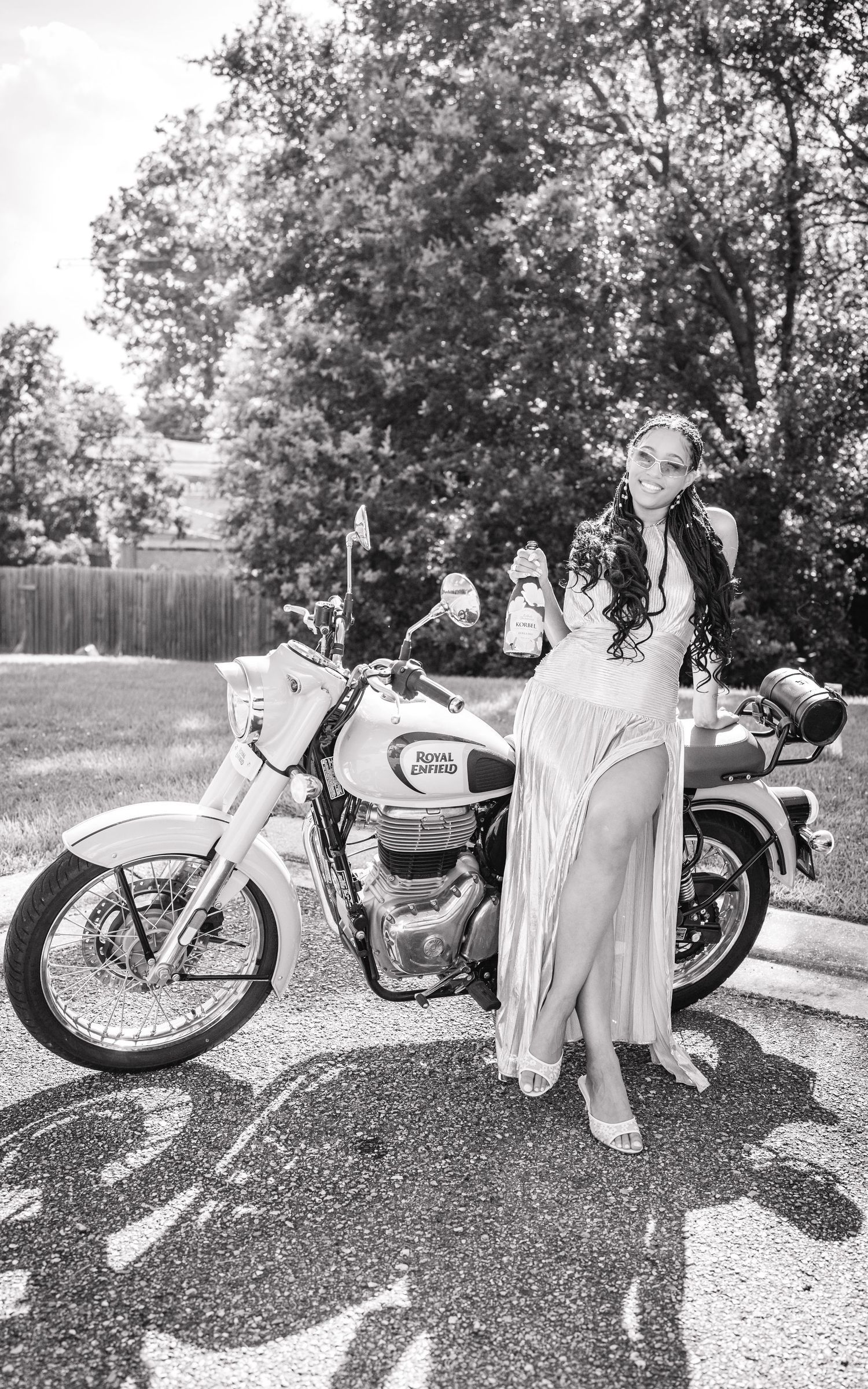 Woman in elegant dress poses beside a Royal Enfield motorcycle on a sunny street in black and white photo.