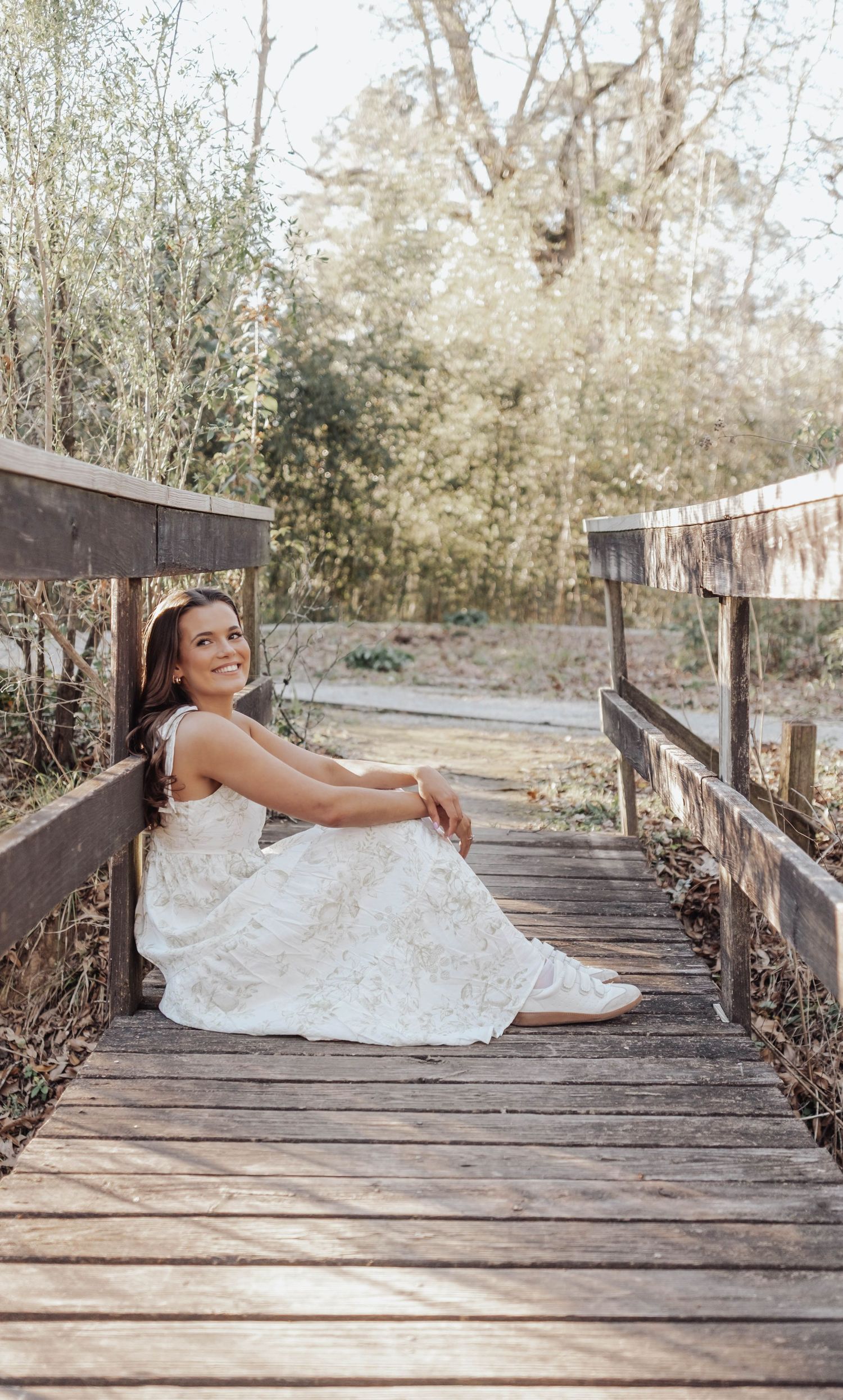 Smiling bride in white lace gown sitting on wooden boardwalk surrounded by lush greenery in natural outdoor setting.