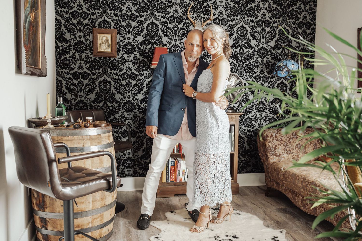 A couple poses intimately against a wall with vintage black and white damask wallpaper in an elegant room with plants.