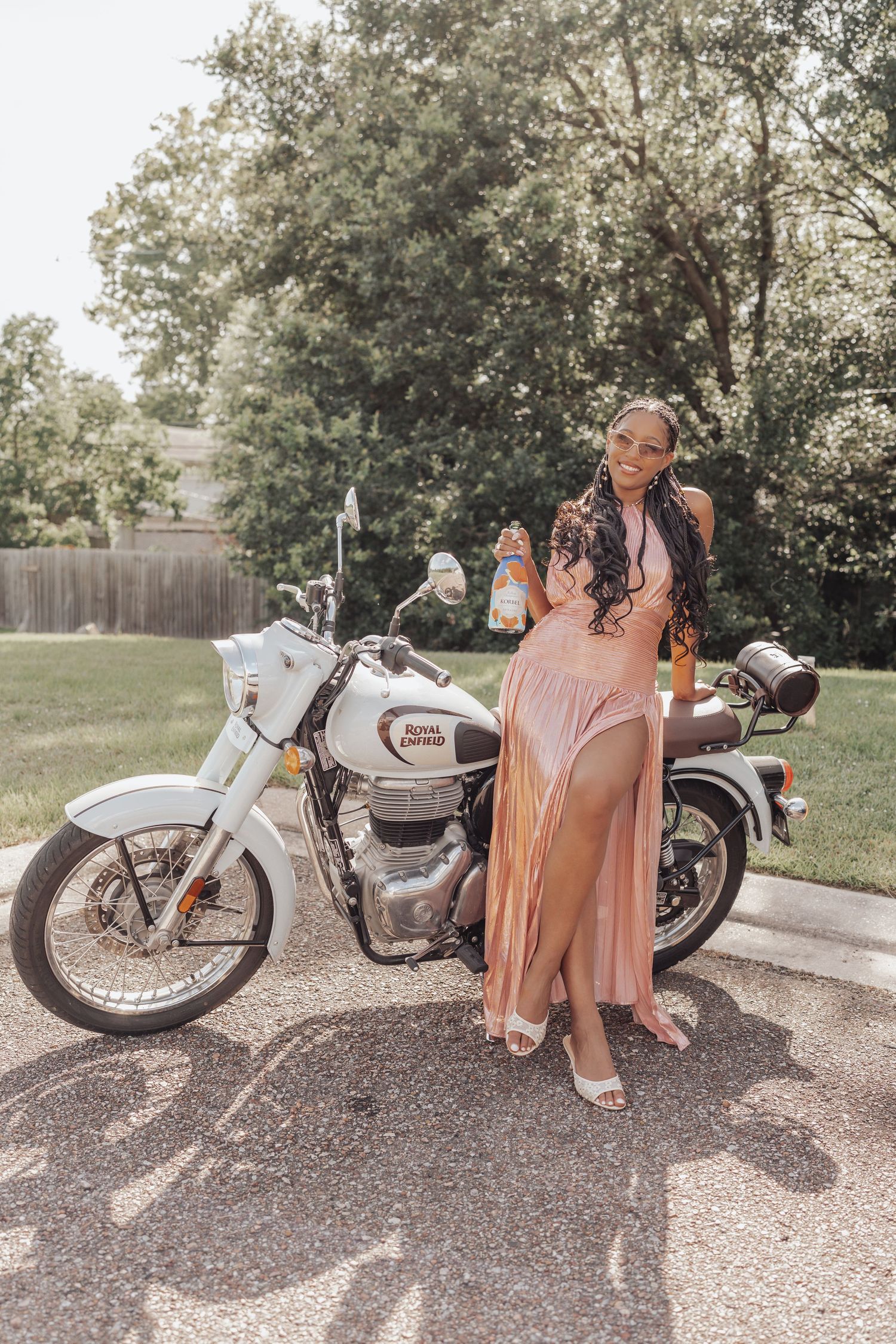 Woman in pink dress posing beside a white Royal Enfield motorcycle in a sunny parking lot.