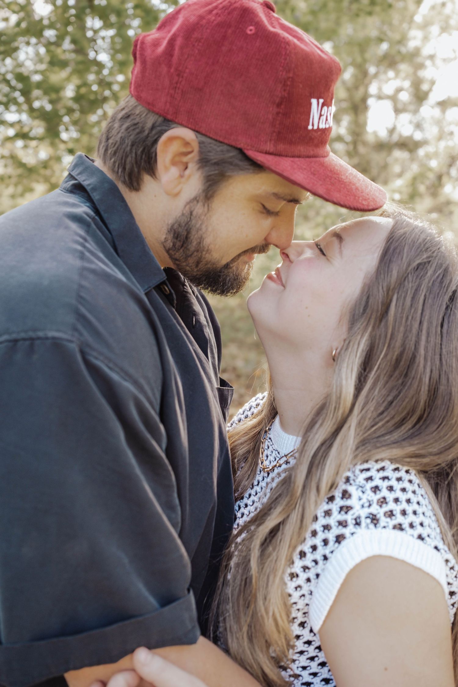 A couple sharing a romantic almost-kiss outdoors, man in red cap and dark shirt, woman in white top, surrounded by trees.