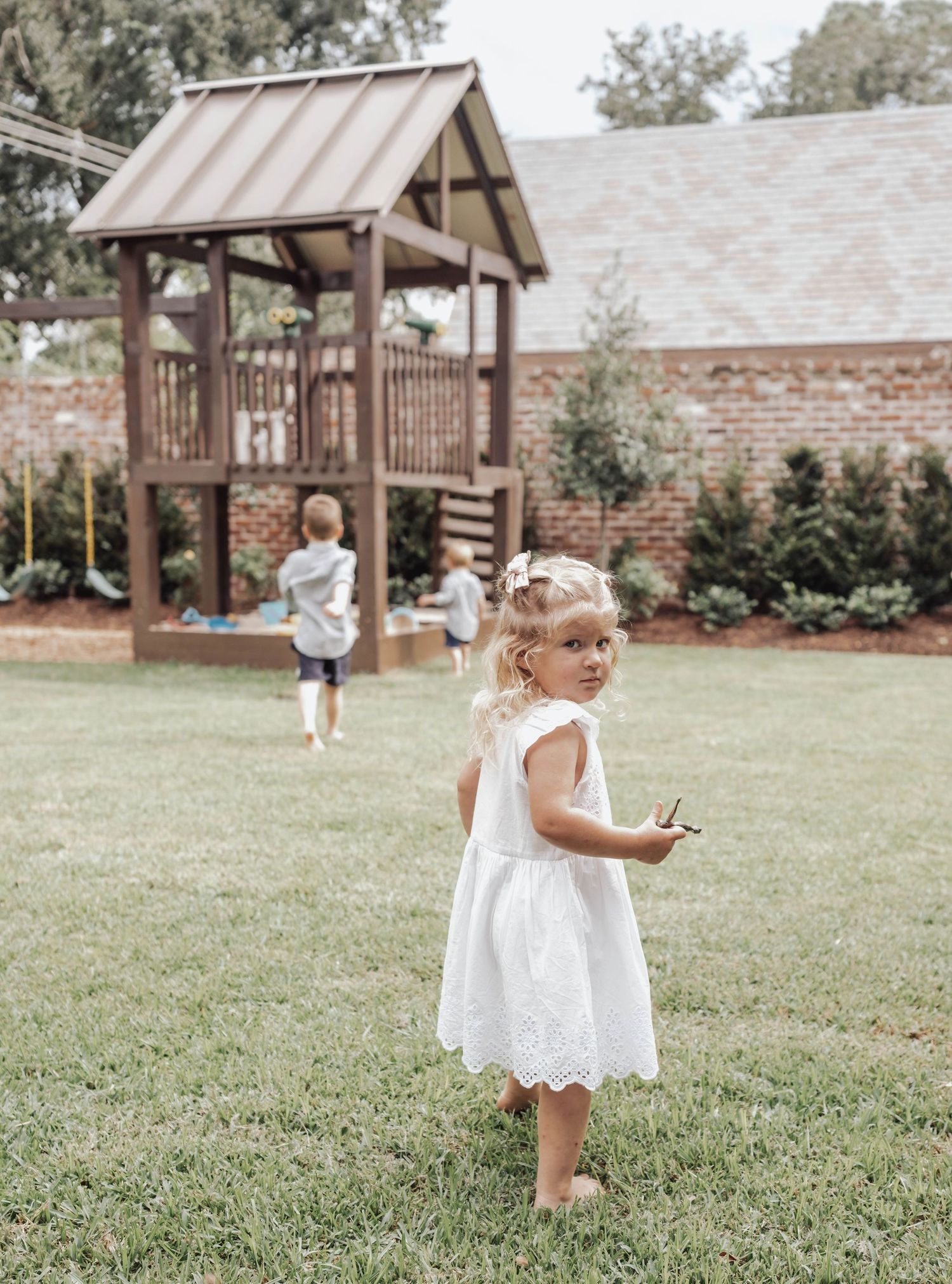 A toddler girl in a white lace dress stands on green grass near a wooden backyard playset.