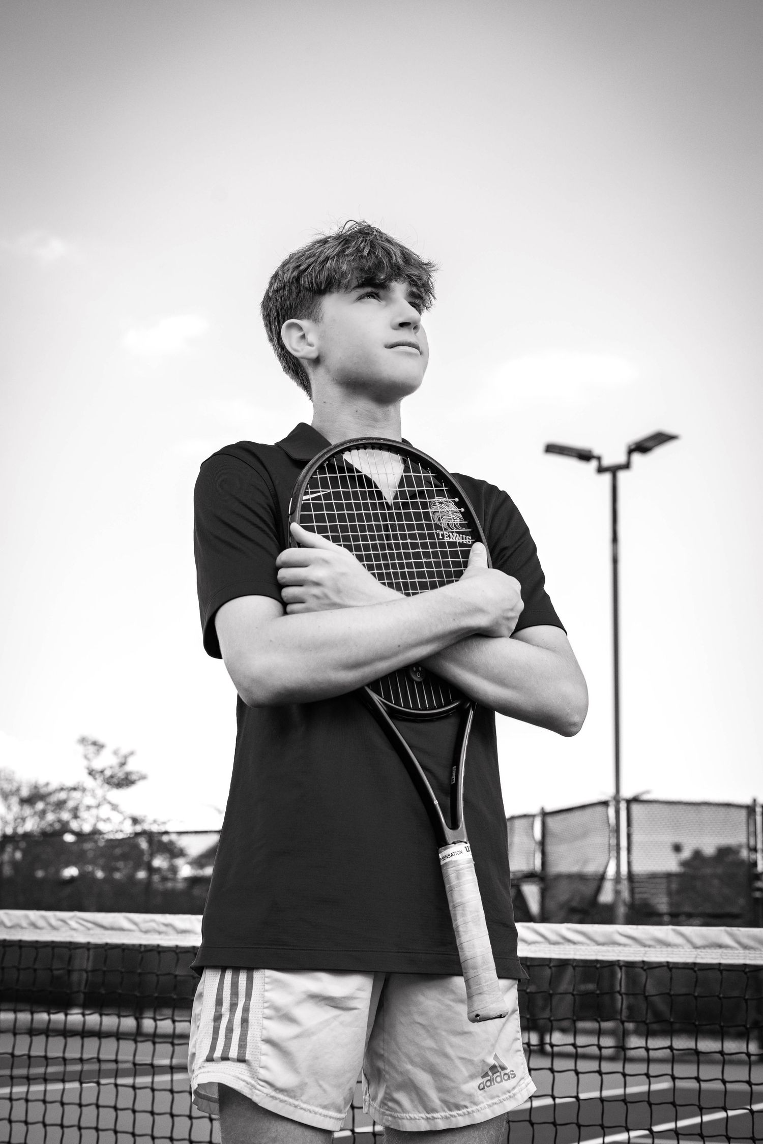 Young tennis player holding racket with arms crossed on court, black and white portrait photo.