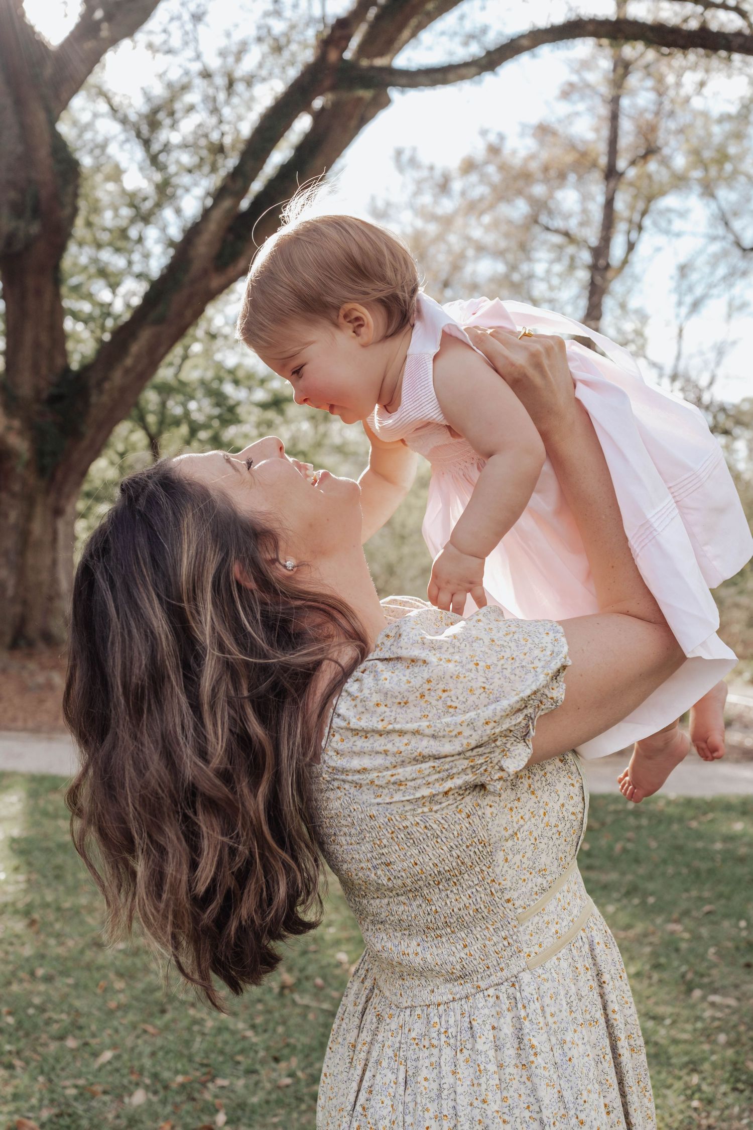 A joyful mother in a floral dress lifts her smiling baby girl in a pink dress outdoors under a tree.