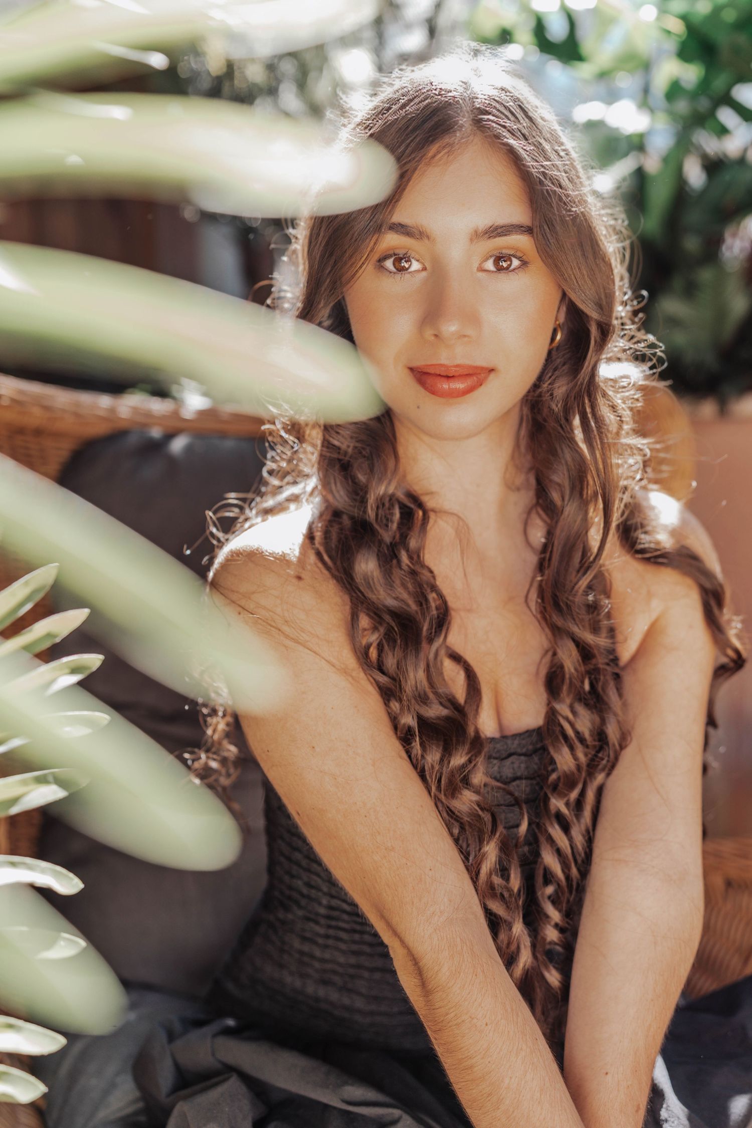 Young woman with long braided hair and red lips poses among tropical green plants in natural sunlight.