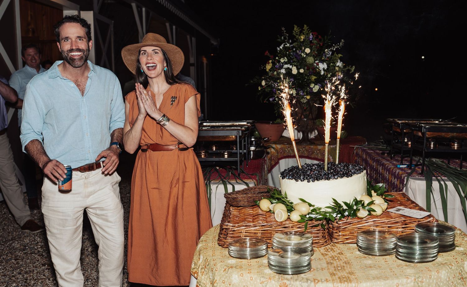 Smiling man and woman in a straw hat celebrate beside a frosted cake with sparklers at an outdoor evening party with decorated tables.