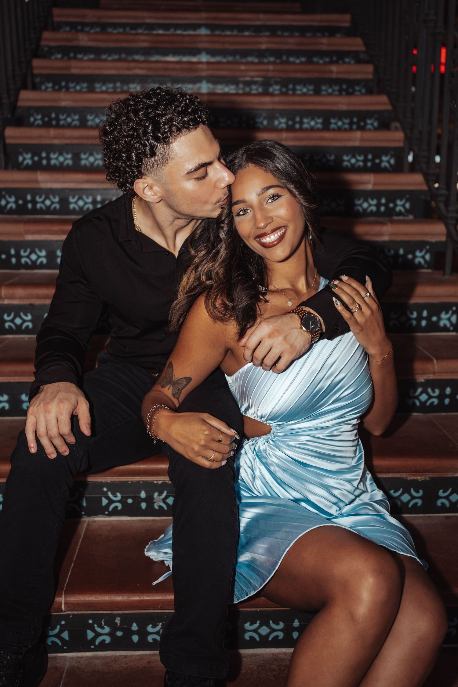 A romantic couple sits on ornate wooden stairs, with one wearing a black shirt and the other in a light blue satin dress.