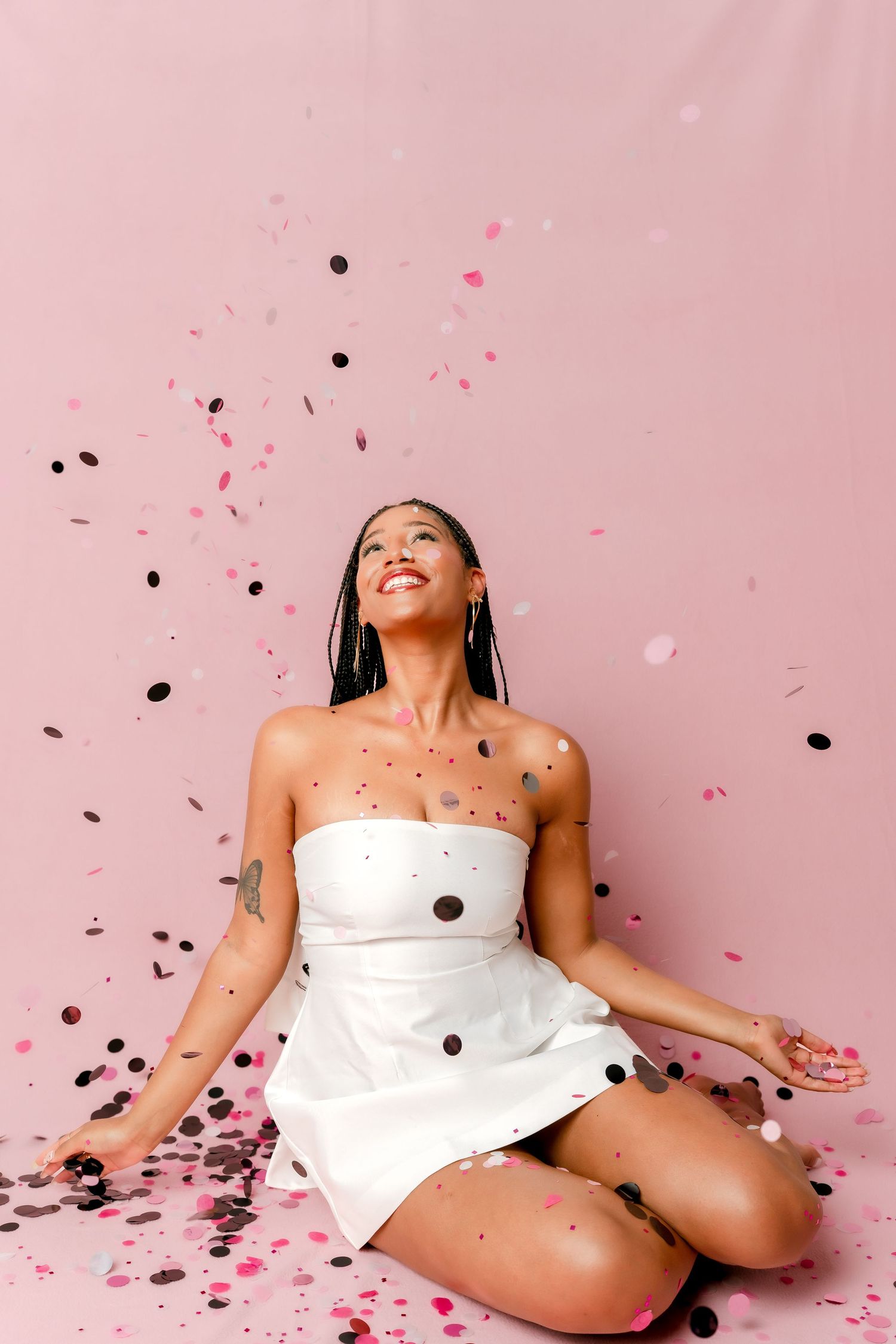 Young woman in white dress sitting on pink floor, smiling up at falling pink and black confetti.