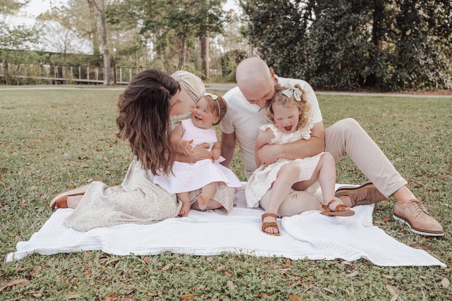 Family of four sitting on white blanket in park, parents and two toddler girls in white outfits sharing a tender moment.