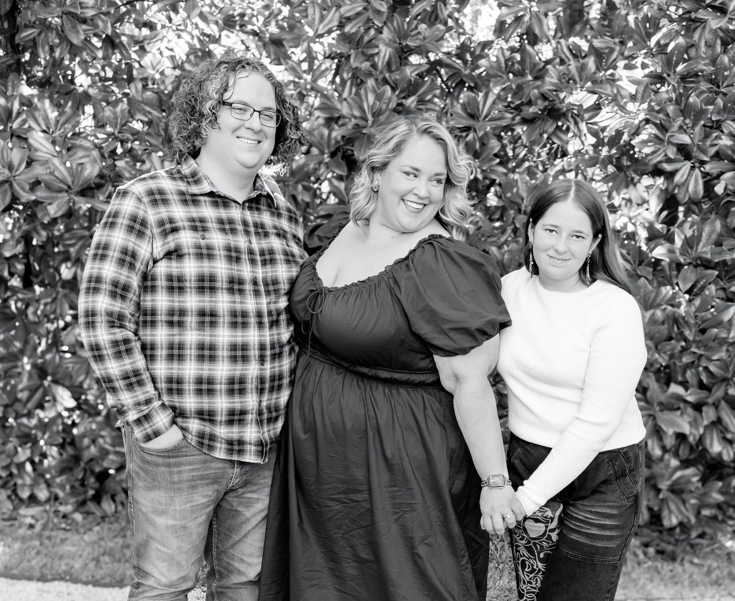 A black and white portrait of three people standing together outdoors against a leafy background.