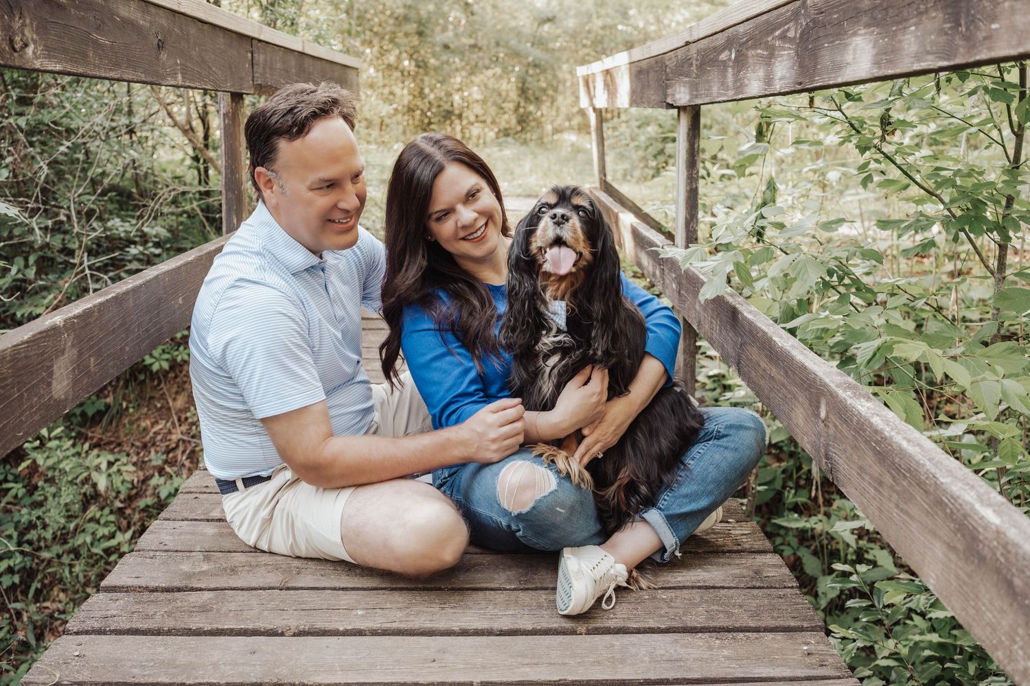 Couple sitting on wooden bridge with black and white Shih Tzu dog surrounded by lush green foliage.