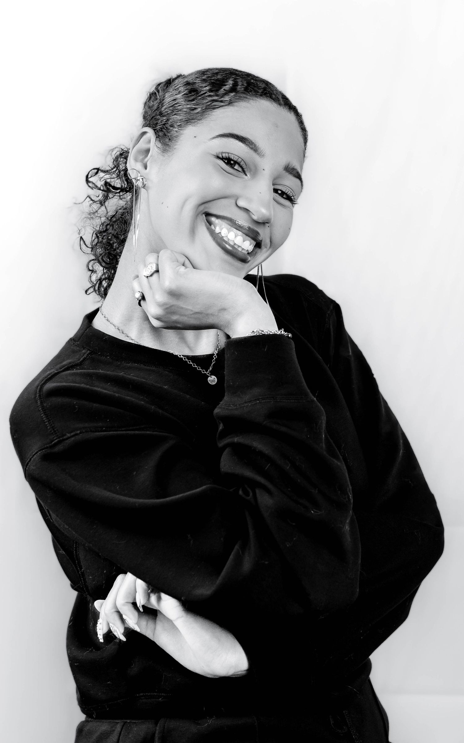 Black and white portrait of a smiling young woman in a black sweatshirt, posing with hand on chin.