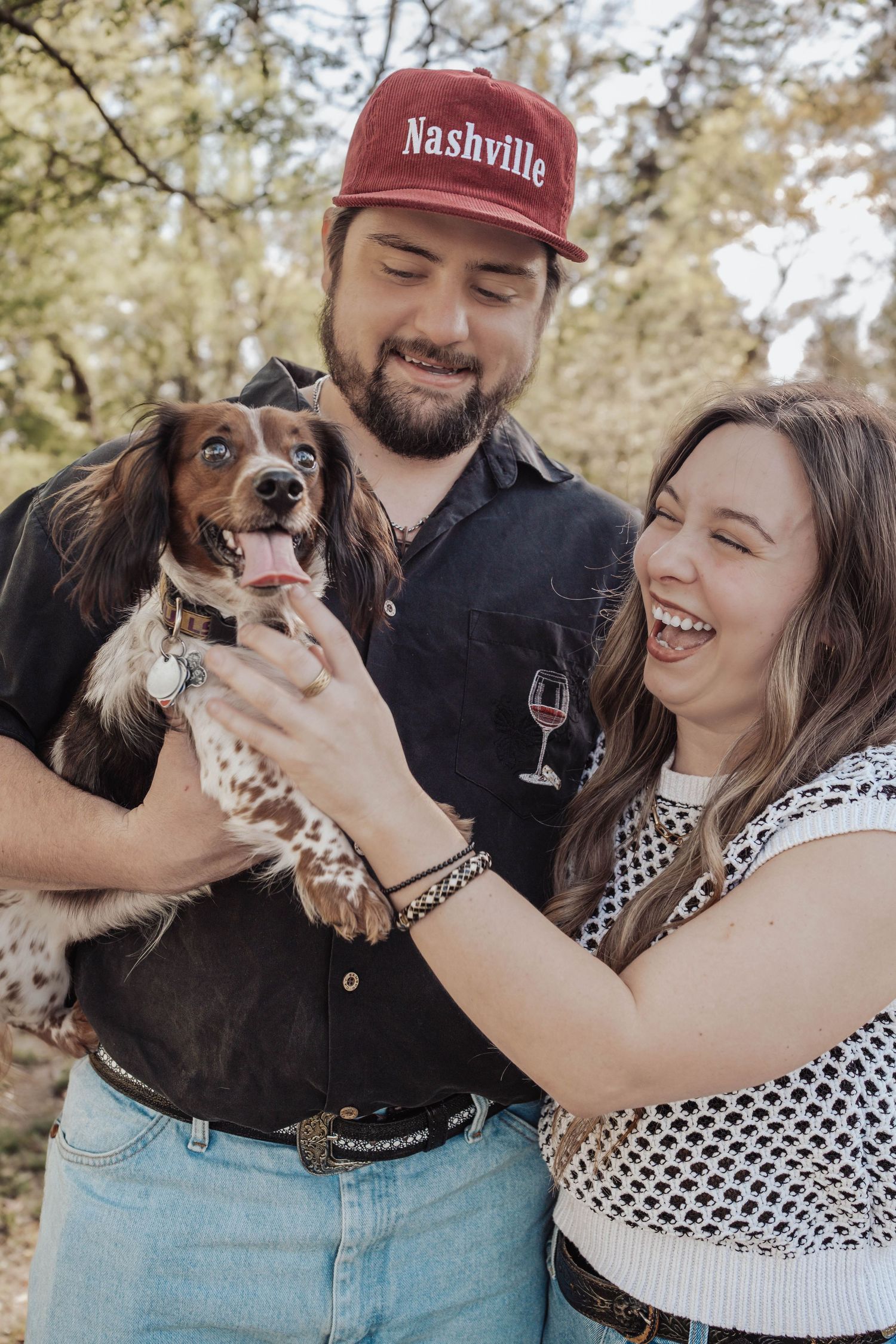 A laughing couple holds an adorable spotted spaniel dog outdoors, man wearing a red Nashville cap and black shirt.