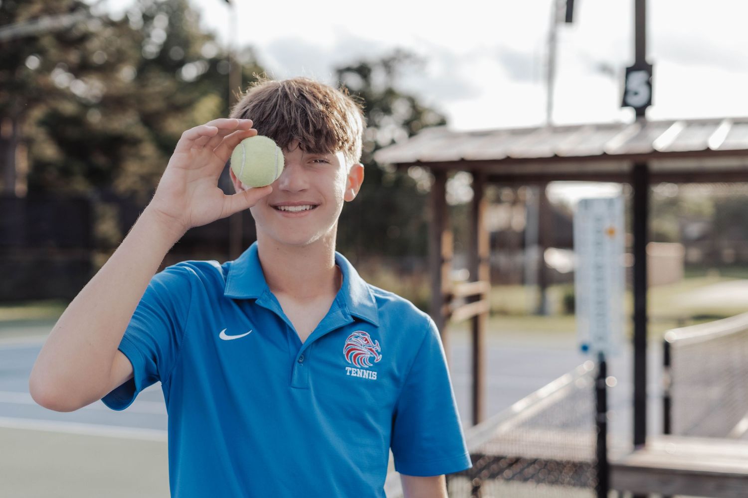 Smiling teenage boy in blue Nike tennis polo holding a tennis ball over his eye on an outdoor court.