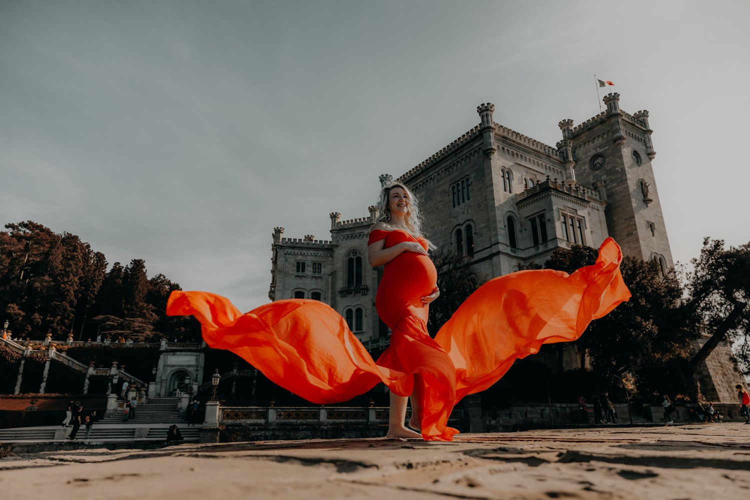 A person in a flowing red dress poses dramatically in front of a gothic building with cloudy skies.