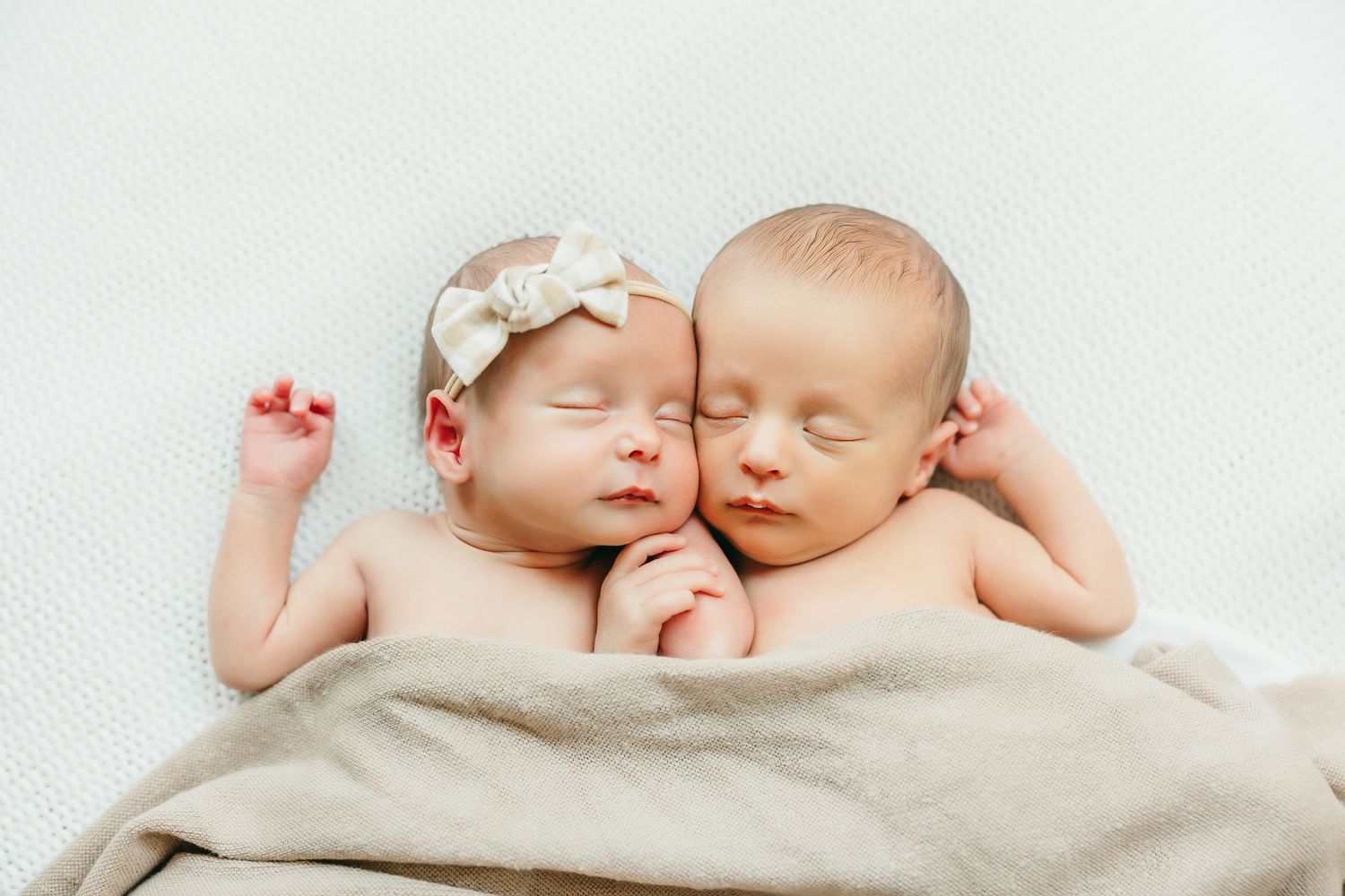 Two sleeping newborn twins snuggled together on a soft blanket, one wearing a bow headband.
