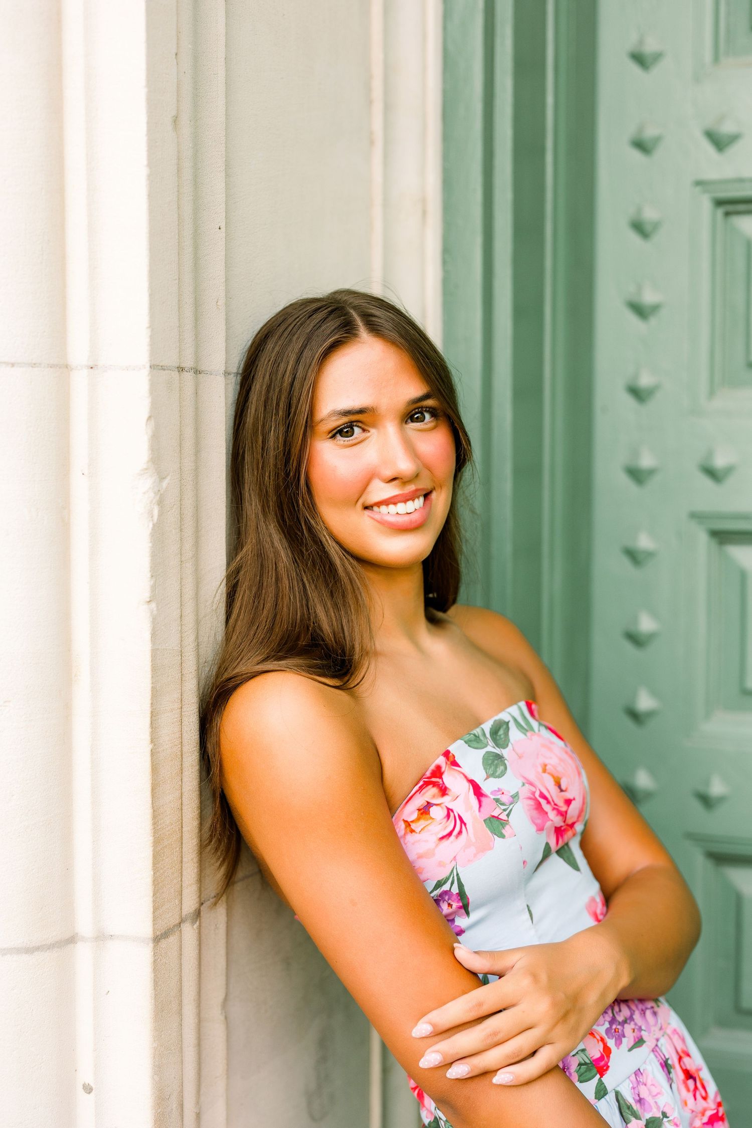 A smiling brunette teen girl in a floral strapless dress leans against a column near a green door.
