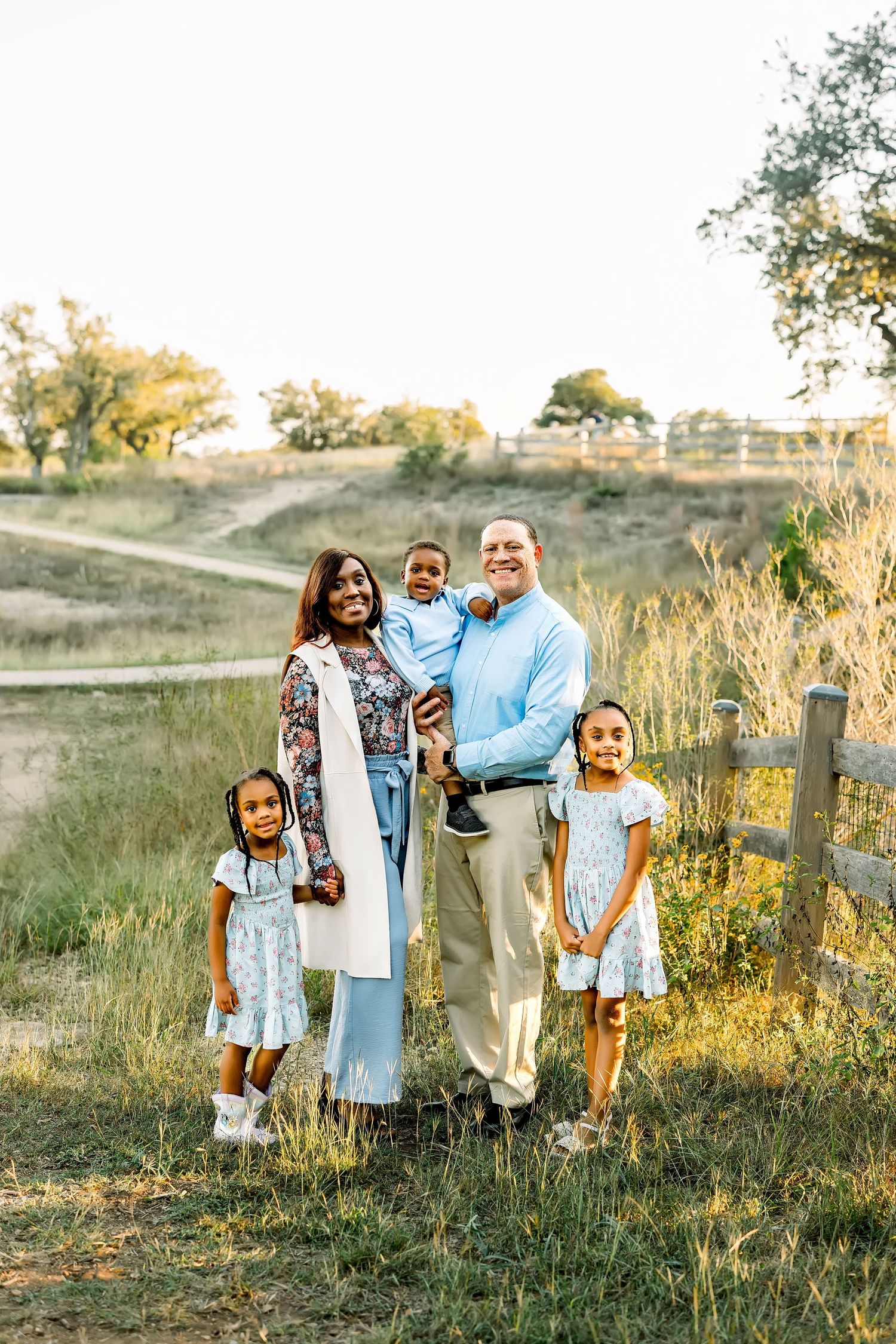 Happy family of five posing near wooden fence in golden hour field setting.