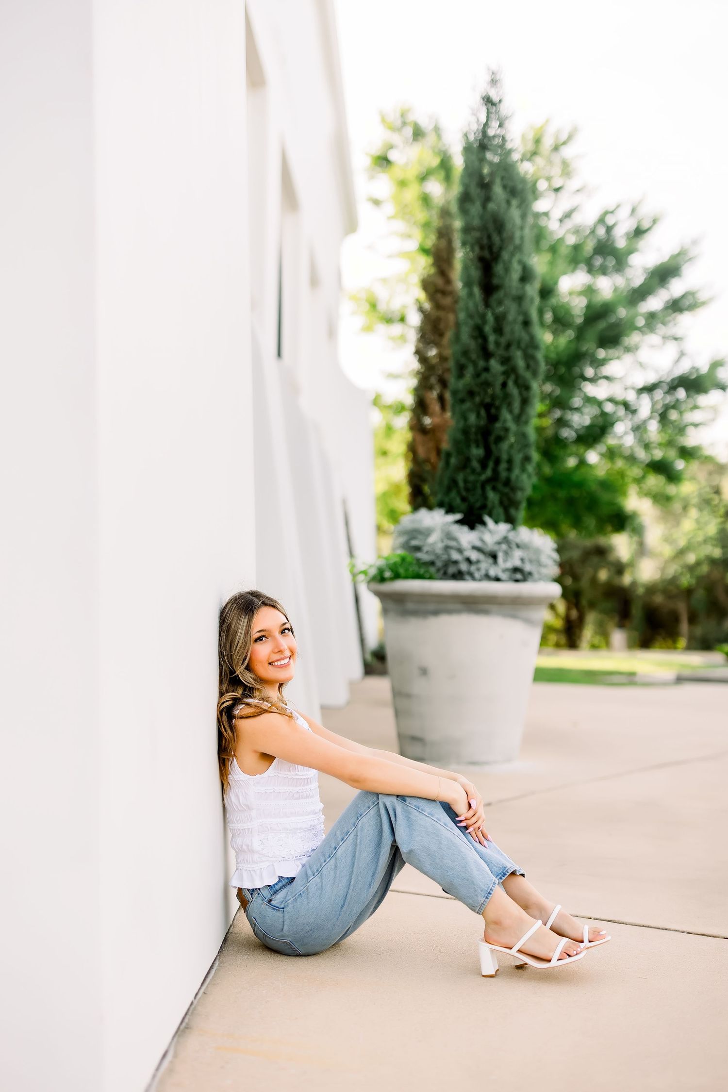 Smiling young woman in white top and jeans sitting against white wall beside potted cypress tree.